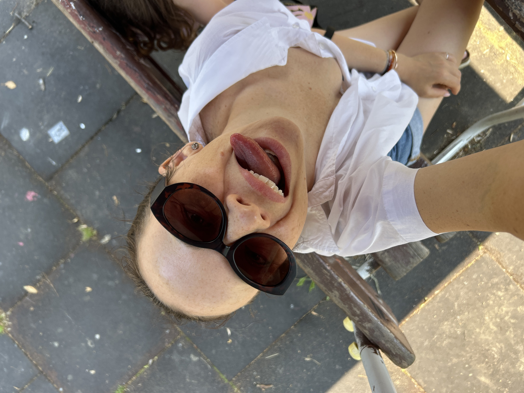A woman is shown from a high angle, appearing to be sitting on a bench outdoors. She is wearing sunglasses and a white shirt, with her tongue sticking out. Her hair is pulled back. The ground appears to be paved with dark gray rectangular stones and there is some grass growing between them. A metal railing is visible in the foreground, and a wooden bench is partially visible beneath her. The image appears to be a selfie taken from an upward-facing perspective. The location is not specified in the image.