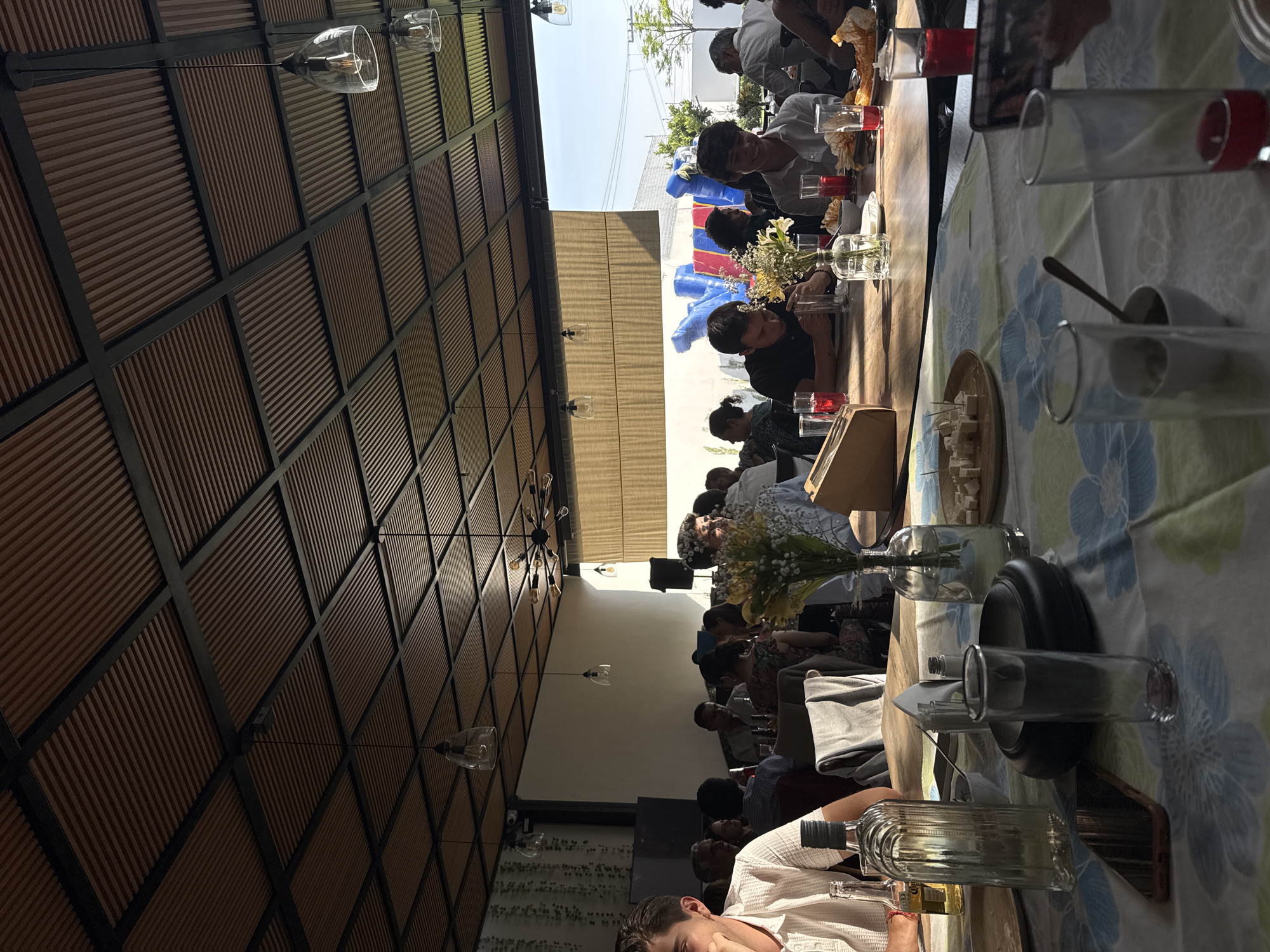 A group of people are seated at a long table in a covered outdoor restaurant. The table is set with flowers, drinks, and food. The restaurant has a distinctive ceiling with a grid pattern and hanging light fixtures. Outside the restaurant, there is a glimpse of blue sky and what appears to be a bouncy castle. The location is San Bernardino Tlaxcalancingo, Mexico.