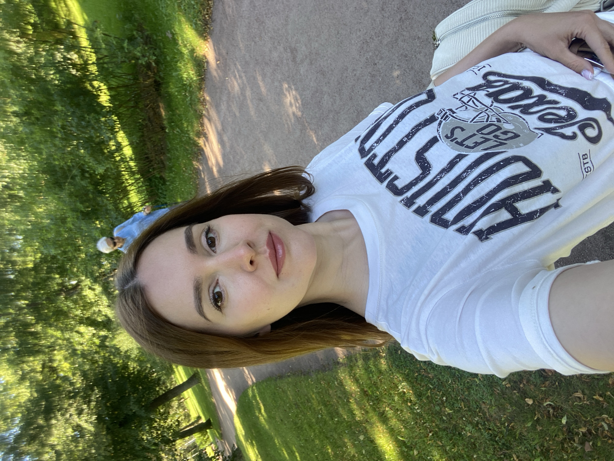 The image shows a young woman taking a selfie in a park. She has brown hair, brown eyes, and is wearing a white t-shirt with "Houston Let's Go Texas" printed on it. In the background, an older woman in a blue jacket is walking away from the camera. The scene appears to be in a park with trees and a path. The location is identified as Peterhof, Russia.