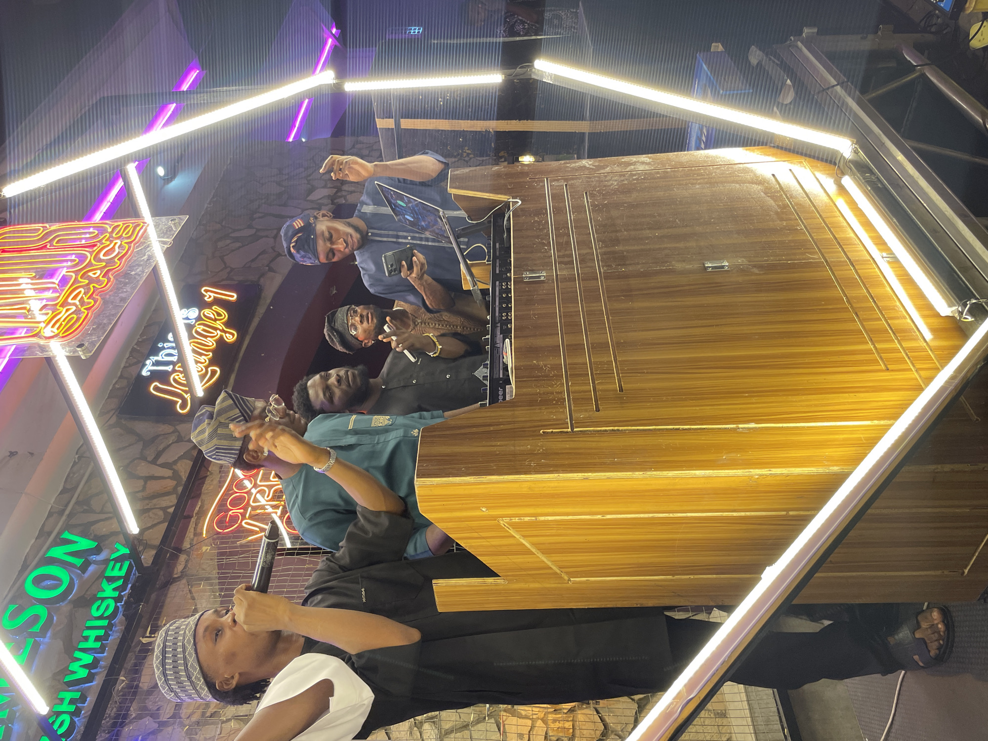 Four men are in a nightclub, performing or recording, in the city of Lokoja, Nigeria. The men are dressed in traditional clothing. One is speaking into a microphone. Another is holding a phone or a tablet. They stand near a dark wooden bar, next to neon signs for Irish Whiskey and "Lounge 1". A frame of white neon lights surrounds them. The background includes a faux stone wall.