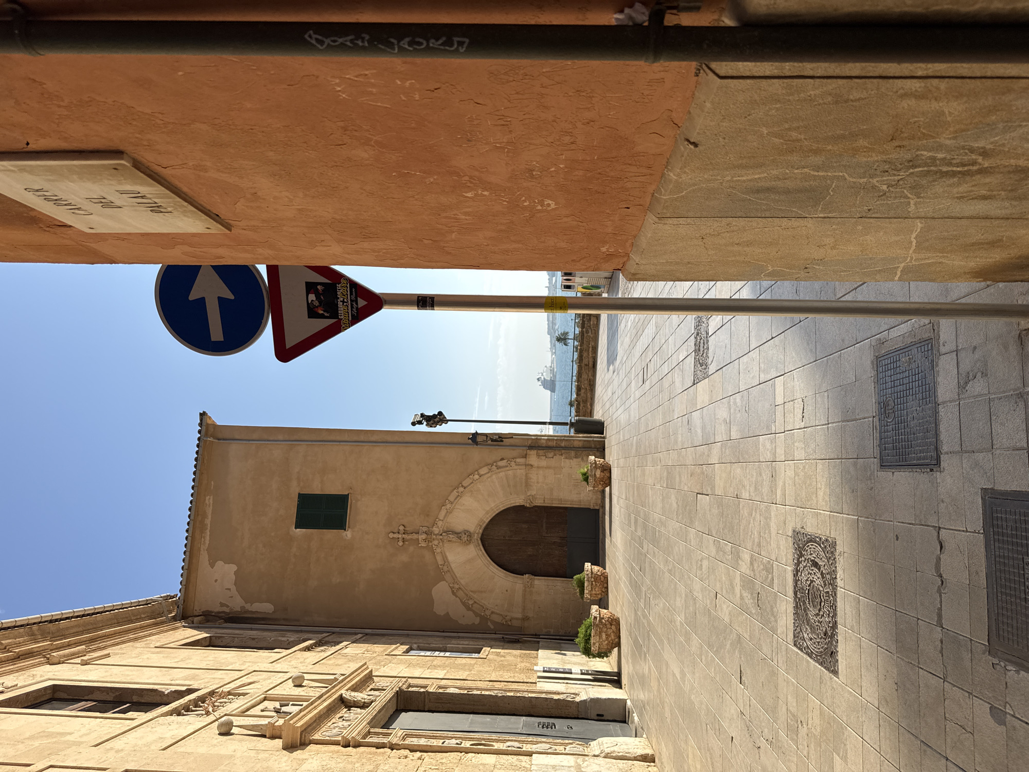 The image depicts a street scene in Palma de Mallorca, Spain. On the left, a building with beige stone facade and intricate carvings around a dark doorway is visible. Three potted plants are placed in front of the building. The street is paved with light-colored square tiles. In the center of the image, a street sign with a blue circular sign indicating a rightward arrow and a triangular yield sign is prominent. Behind the signs, the sea and a large ship are visible. On the right, a portion of an orange building wall is shown with graffiti and a metal pipe running vertically. The sky is clear and blue.
