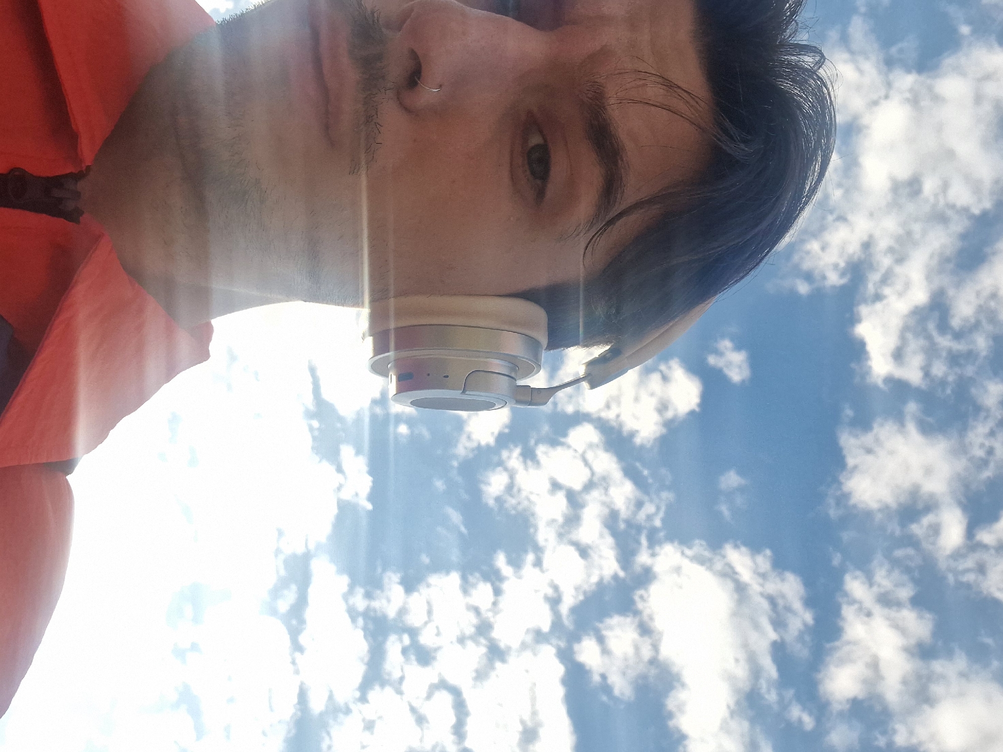 A low-angle, upward-facing view shows a person's face, angled towards the viewer and partially in the frame, with a blue sky and white clouds in the background. The person has dark hair, a mustache, and a nose ring. They are wearing headphones, with the right earcup visible. The person is also wearing an orange shirt. The image is taken in a location that cannot be confirmed as the city of Unknown, Unknown based on the visual data.
