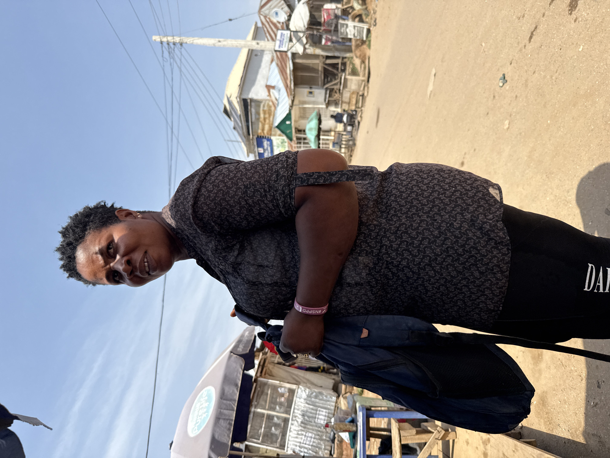 A woman is standing on a dirt road in Abuja, Nigeria, holding a dark blue backpack. She is looking to her left, with her mouth slightly open. She has short, dark, curly hair and is wearing a dark patterned shirt and black leggings. A pink bracelet with white text is visible on her right wrist. Behind her, there are several small shops and market stalls lining the road. The sky is clear and blue, with a few scattered clouds. The ground is dusty and dry, with some patches of water. A power pole with many wires is visible in the background.