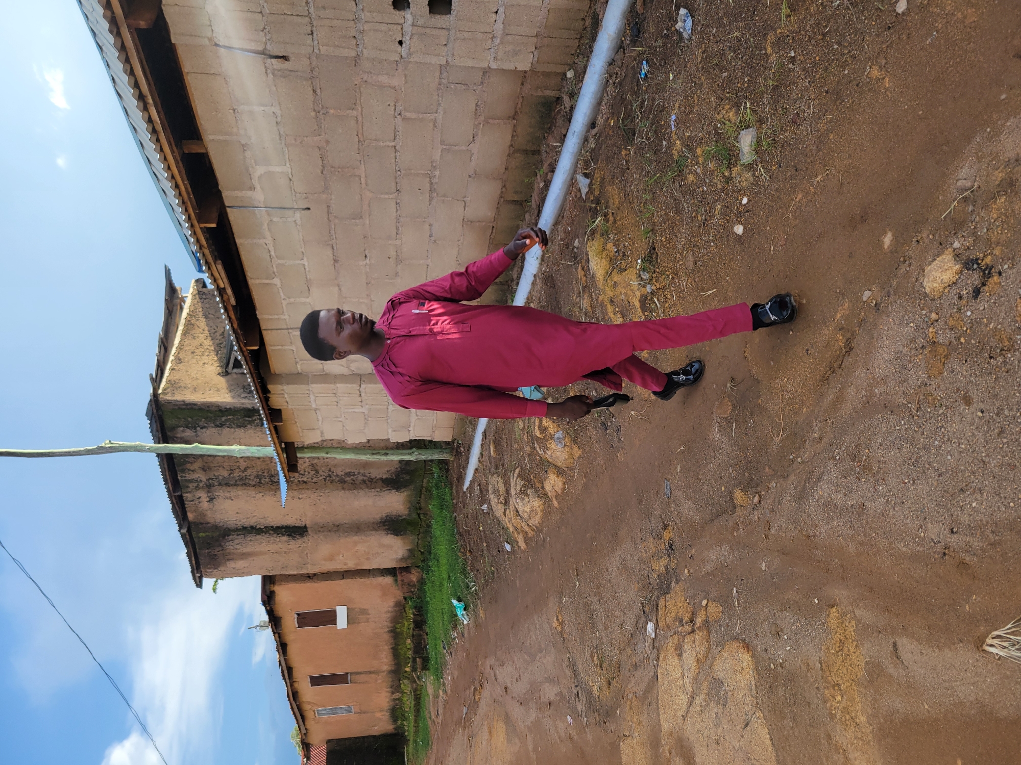 In Jos, Nigeria, a young man in a maroon traditional outfit walks along a dirt road. The road is flanked by buildings. To the left, there are single-story structures with brown, uneven walls and corrugated metal roofs. One building has three visible windows, and another appears to have two. To the right, a building constructed with light-colored concrete blocks has a corrugated metal roof. A thin, pale green pole stands vertically in front of this building. The ground is a mixture of dirt, sand, and scattered rocks, with some sparse green vegetation and debris present. The sky above is blue with scattered white clouds. The man is walking from left to right in the frame, holding a dark object in his right hand. He is wearing black shoes.