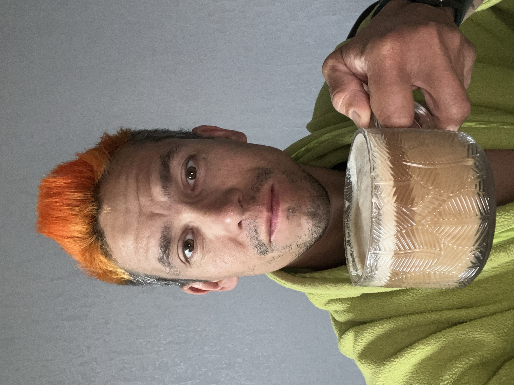 An indoor, close-up portrait shows a man looking directly at the camera while holding a clear glass mug. The man has short, spiky hair, vibrantly dyed orange on top and transitioning to a natural gray or darker color on the sides. He has light stubble covering his chin and upper lip. His brown eyes are open wide, and his expression is neutral to slightly serious. He is wearing a lime green, possibly fleece, hooded garment.
His right hand holds a transparent glass mug that features a distinctive textured pattern of leaves or feathers on its exterior. The mug contains a light-colored, frothy beverage, possibly a milky coffee or a similar drink. A dark wristband or watch is visible on his left wrist. The background is a plain, light gray surface, indicating an indoor setting. There is no visible text in the image. The man appears to be posing for a direct shot or selfie. This scene takes place in Chaikovsky, Russia.