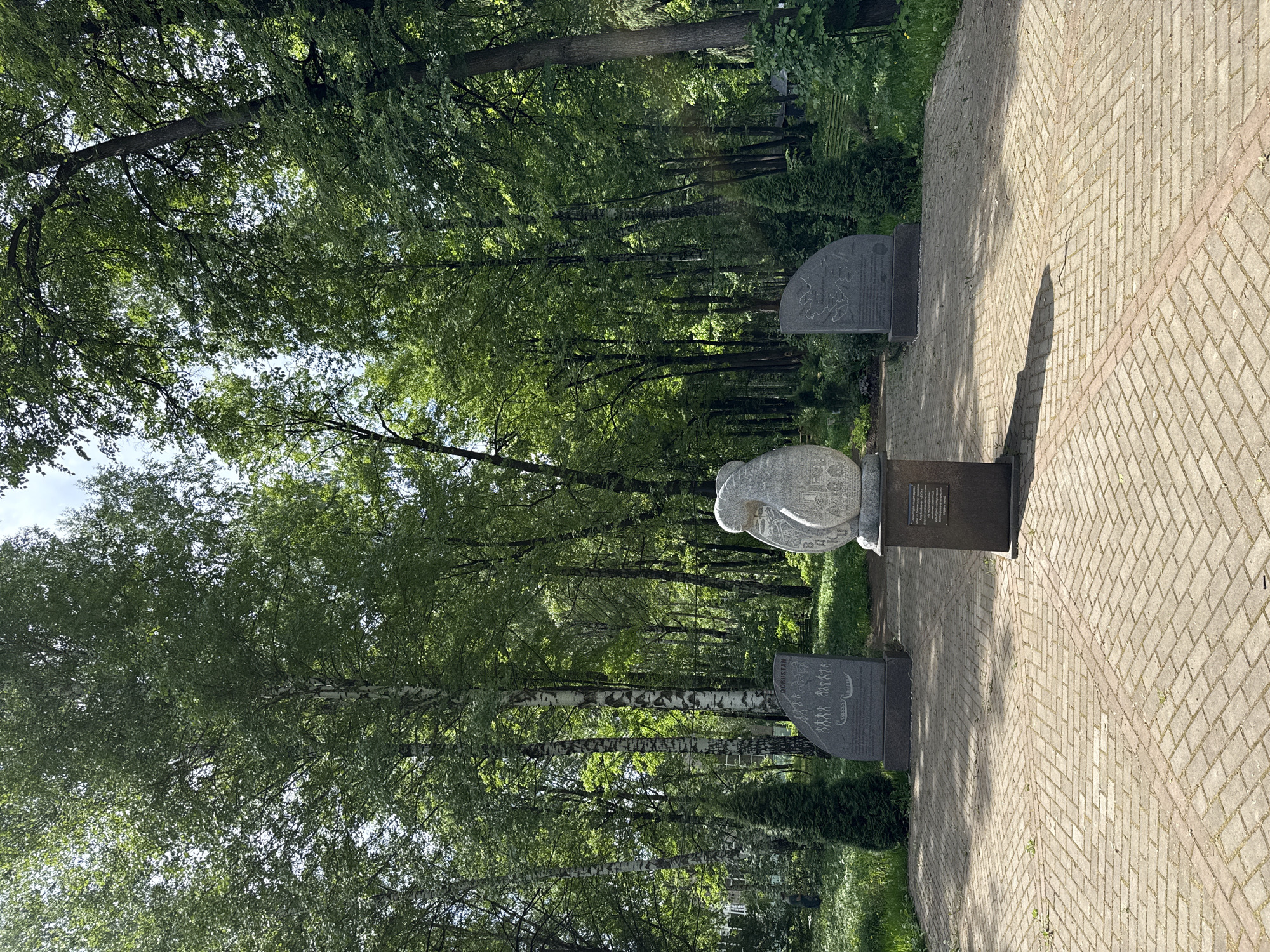 The image shows a stone sculpture and memorial stones along a brick pathway in a park-like setting with numerous trees. The location is Izhevsk, Russia. The sculpture is a rounded form with etched designs, placed on a dark metal or stone stand. Two memorial stones flank the sculpture along the pathway. The pathway itself is made of light-colored bricks laid in a herringbone pattern. The trees behind the memorials are predominantly green and leafy. The scene is well-lit, suggesting a sunny day. The image appears to be taken at an angle, rotating the objects clockwise.