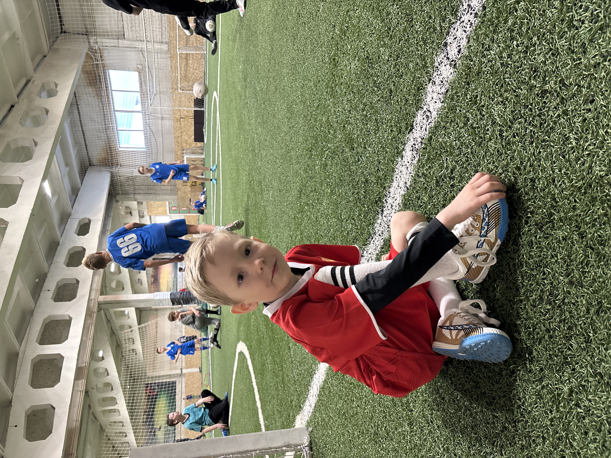 A young child in a red soccer uniform is sitting on an indoor soccer field, close to the white sideline. The child is wearing white socks and soccer shoes with light blue soles. Other children in blue soccer uniforms are playing in the background. The indoor soccer field is likely located in the city of Chaikovsky, Russia. The environment includes the soccer field with artificial turf, and a netting system, likely to prevent balls from leaving the playing area. Part of a goal is visible in the foreground. The scene is brightly lit.