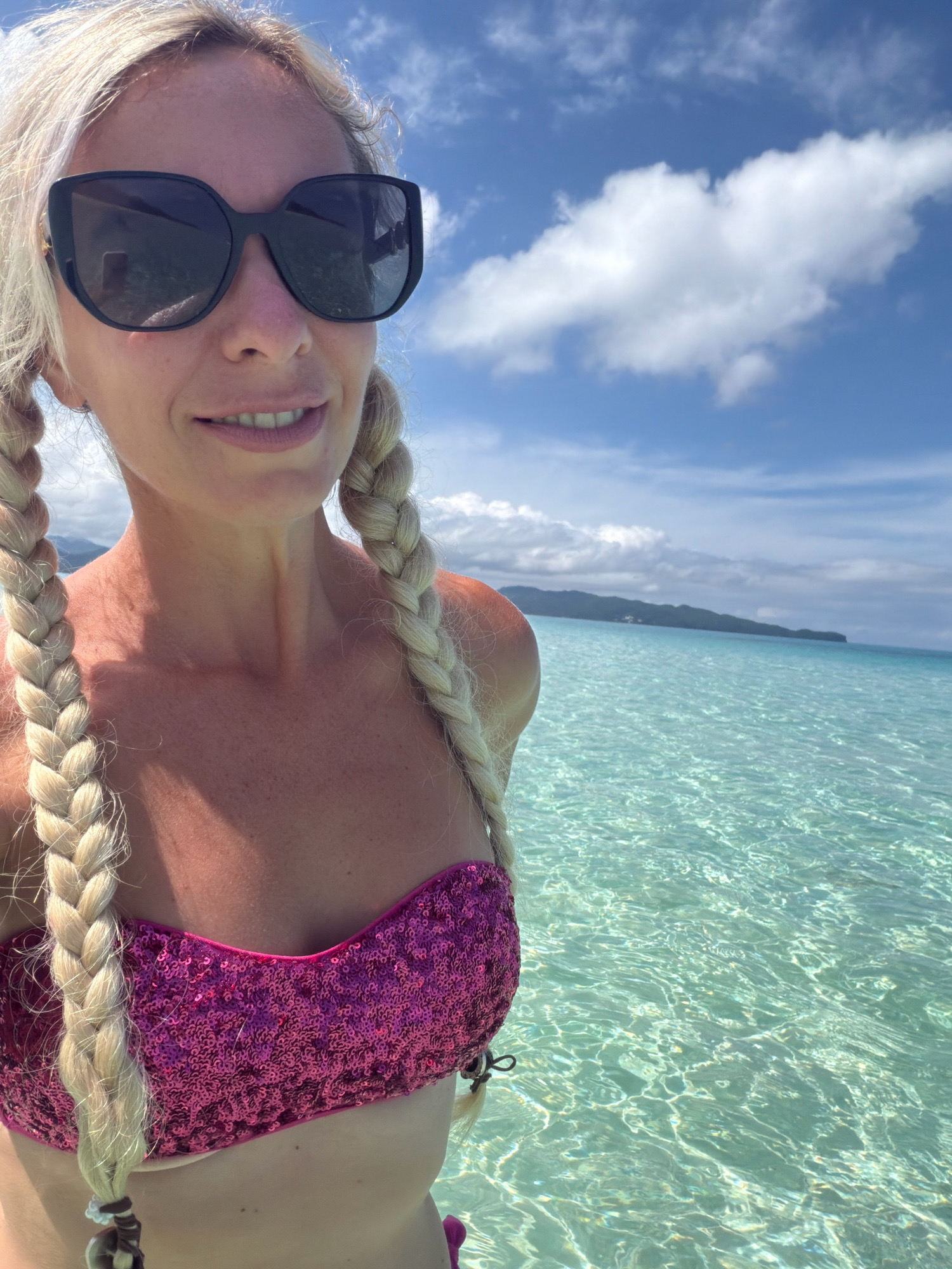 A woman with blonde hair in braids and a pink sequined bikini top stands in shallow, clear turquoise water. She is wearing large black sunglasses and a slight smile. The background shows a bright blue sky with white clouds, a distant green island, and the glistening surface of the ocean. The location is likely a beach in Malay, Philippines, given the characteristic clear waters and island scenery.