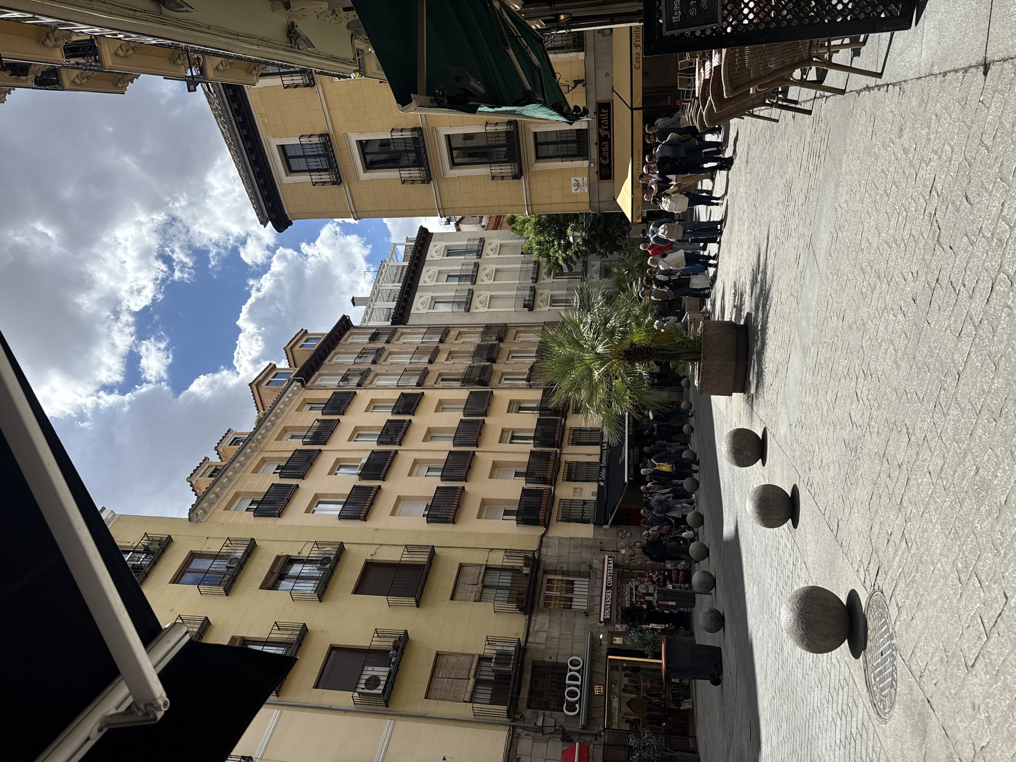 The image shows a street scene in Madrid, Spain. The buildings are mostly yellow stucco with dark wrought iron balconies. A palm tree is centrally placed on the street. There are a number of people standing in a line. On the street itself, there are decorative round stone bollards placed to prevent vehicle access. The sky is partly cloudy with patches of blue sky. There are signs for businesses, including "Casa Fraile", "CODO", and "SOUVENIRS CONTRERAS."
