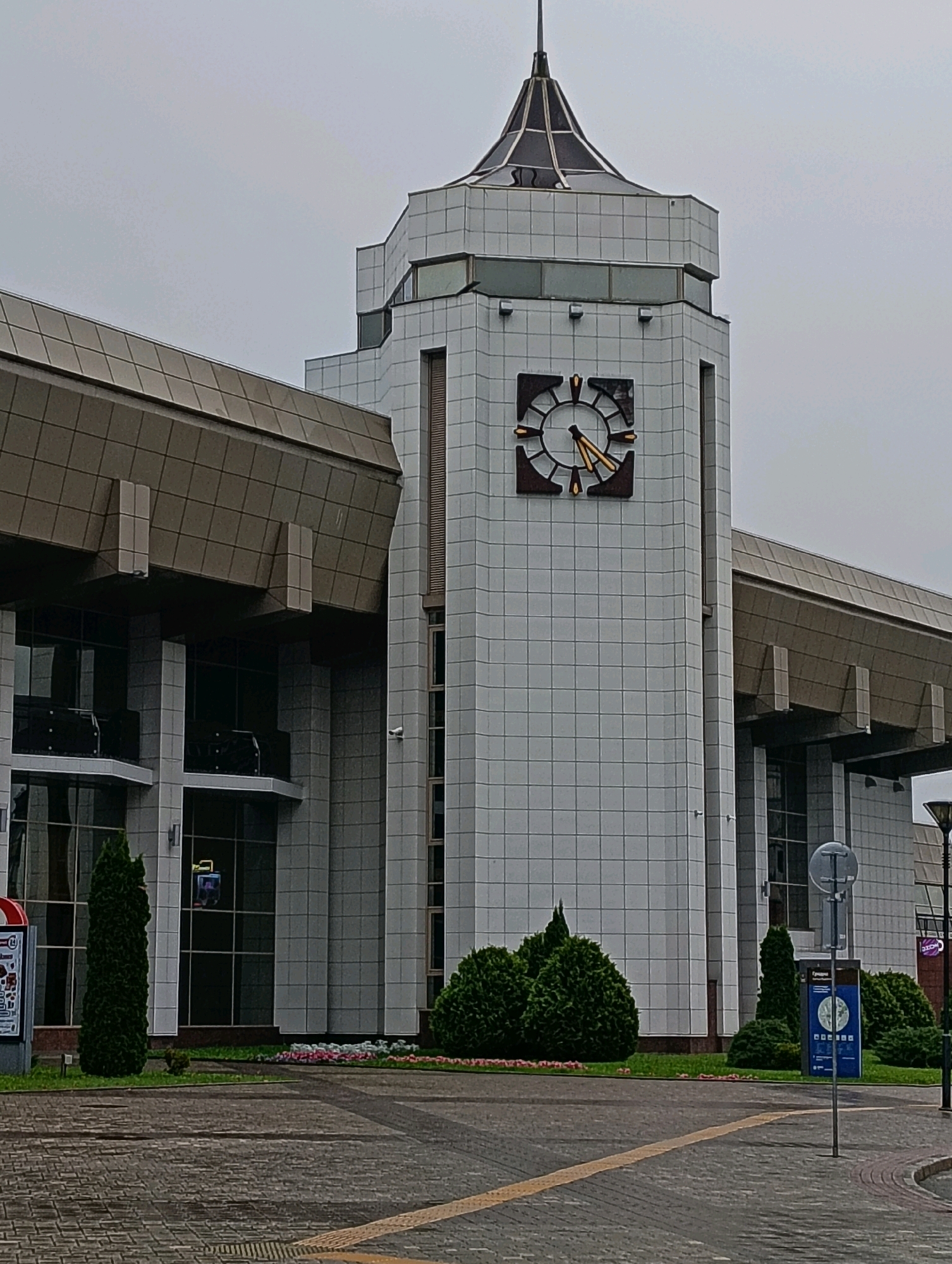 The image shows the Hrodna railway station in Hrodna, Belarus. The station has a modern architectural style with a prominent clock tower. The building is clad in white tiles and features large glass windows. In front of the station, there is a paved area with some landscaping, including bushes and flower beds. The sky is overcast. There are no people visible in the image.