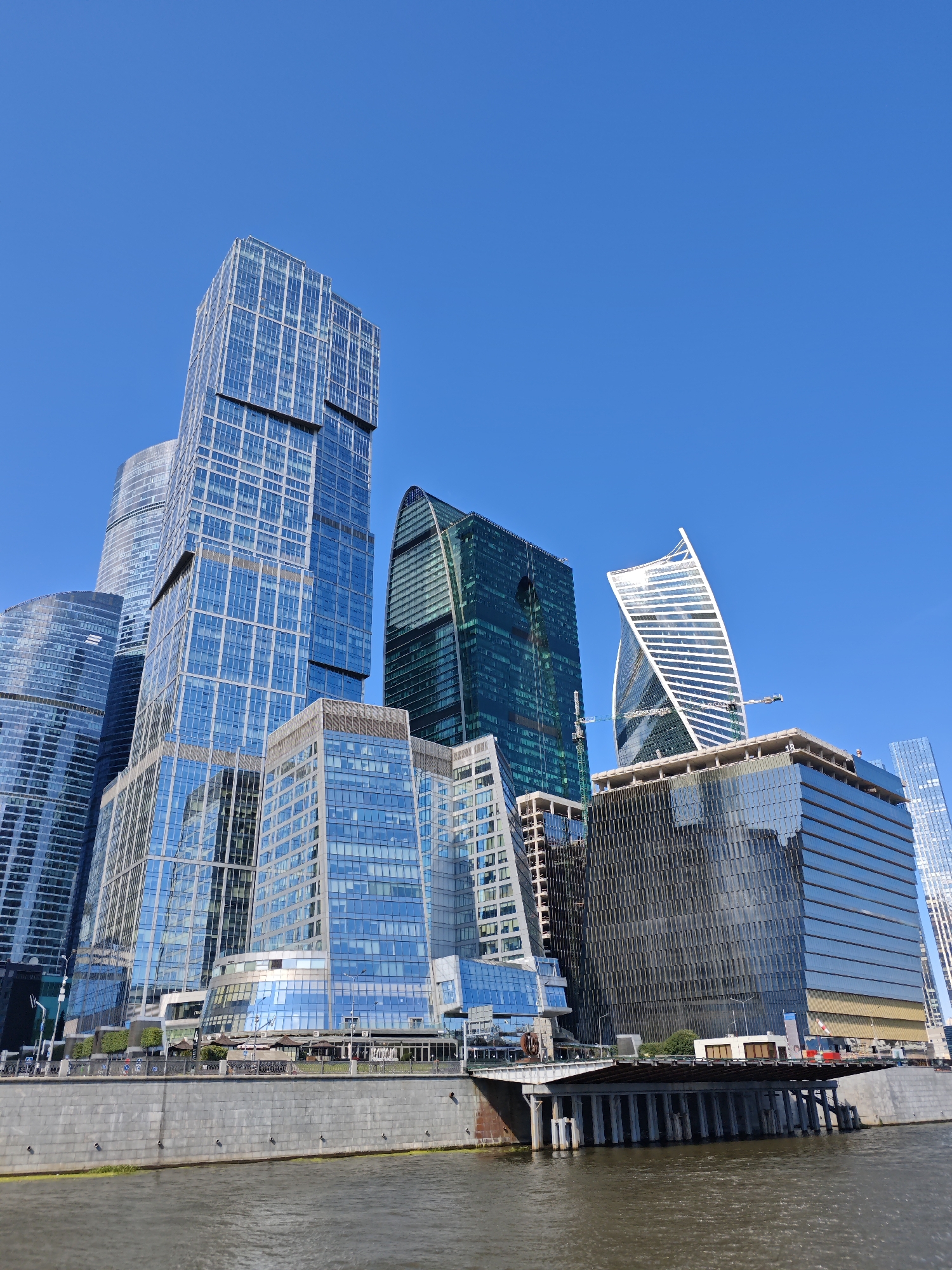 The image depicts a view of the Moscow International Business Center, also known as Moscow-City, a commercial district in Moscow, Russia. Several modern skyscrapers with glass facades are prominently featured against a clear blue sky. In the foreground, a body of water, likely the Moskva River, separates the viewer from the embankment. The embankment itself is lined with a stone wall and a railing. Beyond the wall, a bridge spans the water, connecting the riverbank to the skyscrapers. The architecture of the buildings varies, with some featuring sharp, angular designs and others displaying more curved, organic forms. One particularly striking building has a twisted, spiraling shape. The scene appears to be a daytime view, with ample sunlight reflecting off the glass surfaces of the buildings. The overall impression is one of a modern, developed urban landscape.