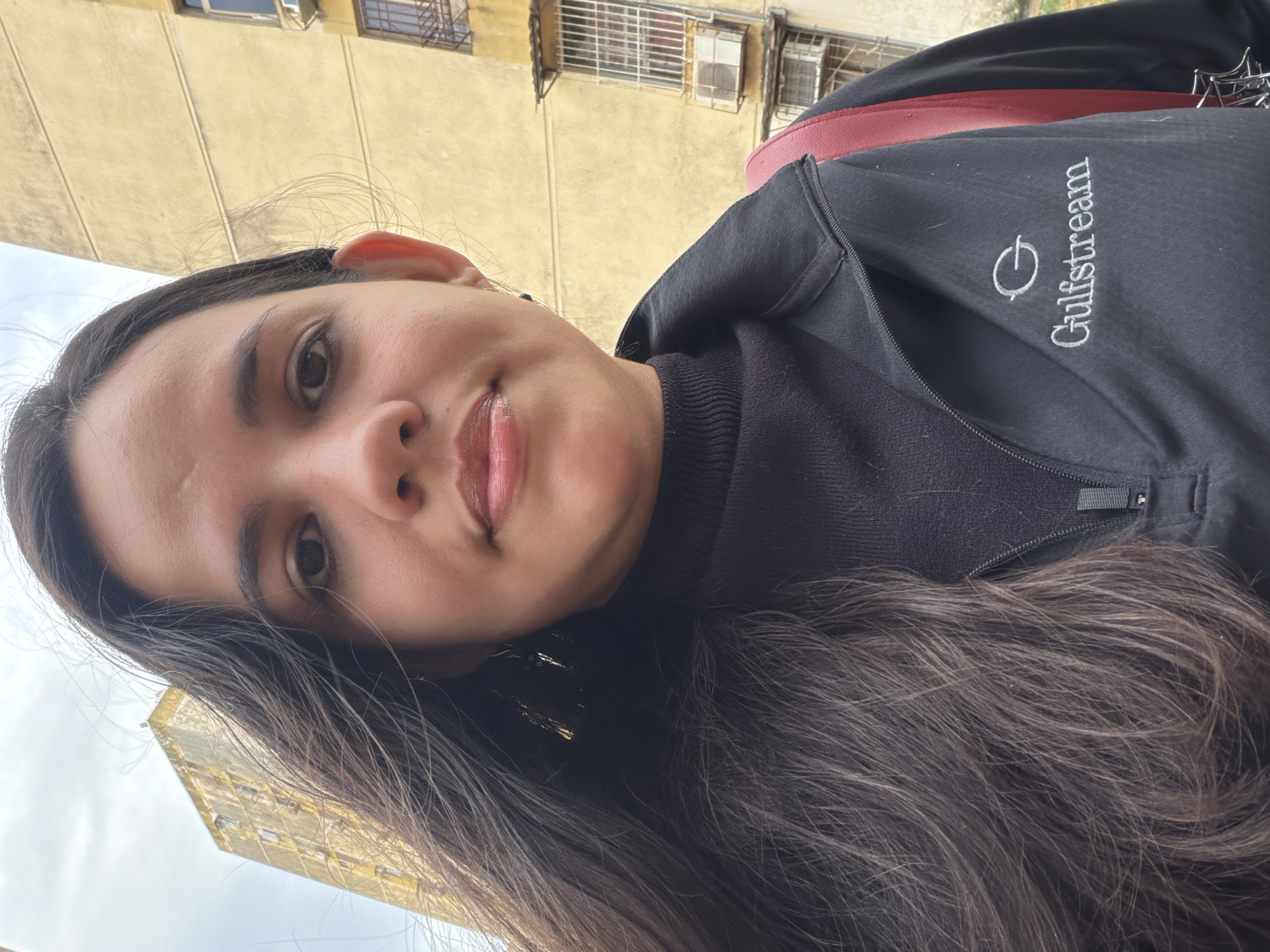 The image depicts a female individual in an urban environment in Ciudad Guayana, Venezuela, posing for a close-up, likely a selfie. She has dark, long, wavy hair, a neutral facial expression, and is looking directly into the camera. She is wearing a black turtleneck sweater and a black zip-up jacket with a white embroidered "Gulfstream" logo on the left chest. A red backpack strap is visible over her right shoulder. In the background, under an overcast sky, are two multi-story buildings, appearing aged with stained yellow facades. The building to the right shows multiple windows, some fitted with metal security bars, and visible external air conditioning units. The overall setting is consistent with a residential or mixed-use urban area.