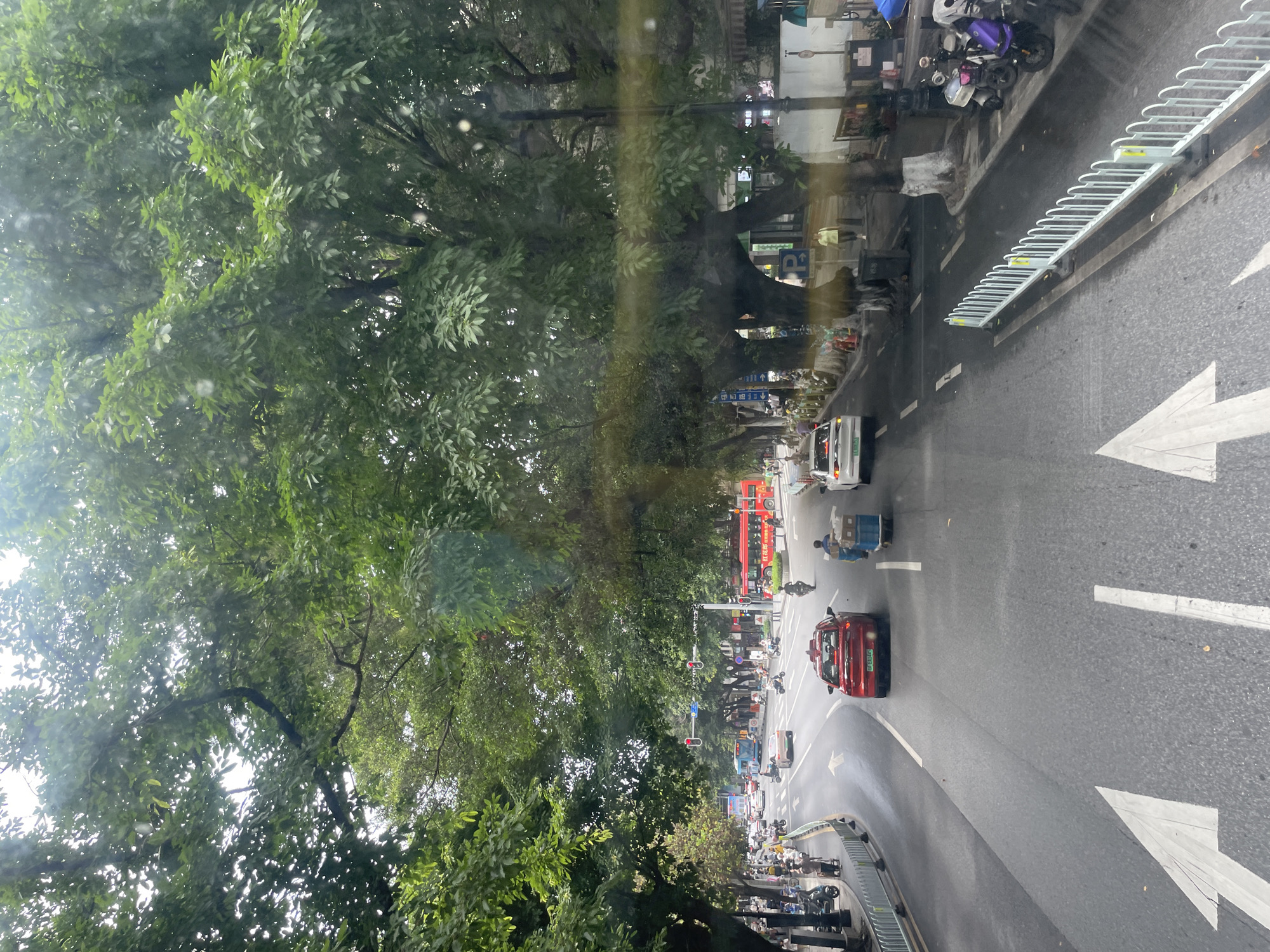 The image captures a street scene in Yuexiu District, China, viewed from an elevated position, possibly a building or a high vehicle. The road is paved and features white directional arrows and lane markings. Traffic includes cars, a red double-decker bus, and smaller three-wheeled vehicles. Pedestrians and parked scooters are visible on the sidewalks. A metal bike rack lines the edge of the sidewalk. Lush green trees overhang the street, creating a canopy effect. In the background, a red traditional Chinese gate is visible amidst the buildings and traffic.