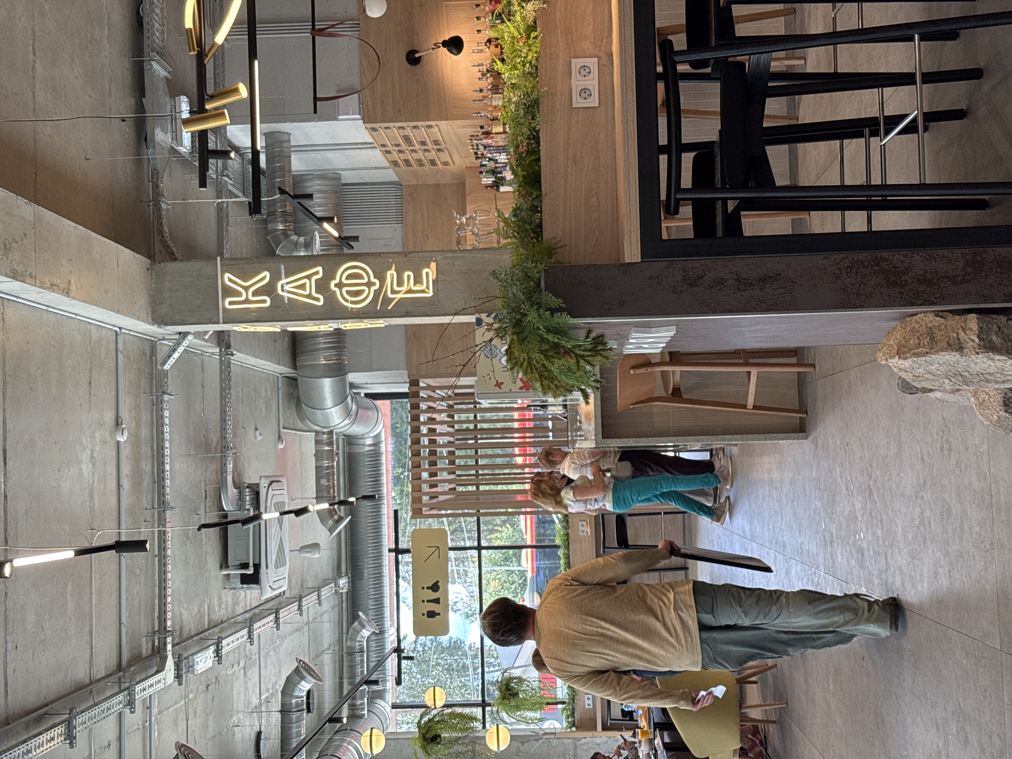 This image depicts the interior of a modern cafe with an industrial aesthetic, characterized by exposed concrete ceilings, ductwork, and lighting fixtures. The cafe appears to be located in Lakhdenpokhya, Russia, as indicated by the Cyrillic neon sign that spells "КАФЕ" (KAFE), meaning "cafe."
In the foreground, a person wearing a tan long-sleeved shirt and green pants walks away from the camera, carrying a dark rectangular object. To their right, two women are standing and conversing, one in a white and tie-dye top and turquoise pants, and the other in a white top and dark pants. They are near a bar area that features a wooden counter, shelves stocked with bottles of alcohol, and an arrangement of fresh greenery.
Behind the people and the bar, a large window offers a view of the outdoors, with trees and a red train visible outside. Above the window, a sign with pictograms indicating restrooms and accessibility is mounted. The cafe's seating includes high stools at the bar and chairs in the background. The overall environment is brightly lit, with a mix of natural light from the window and artificial lighting from various fixtures on the ceiling and walls. The floor is tiled with large gray tiles.
