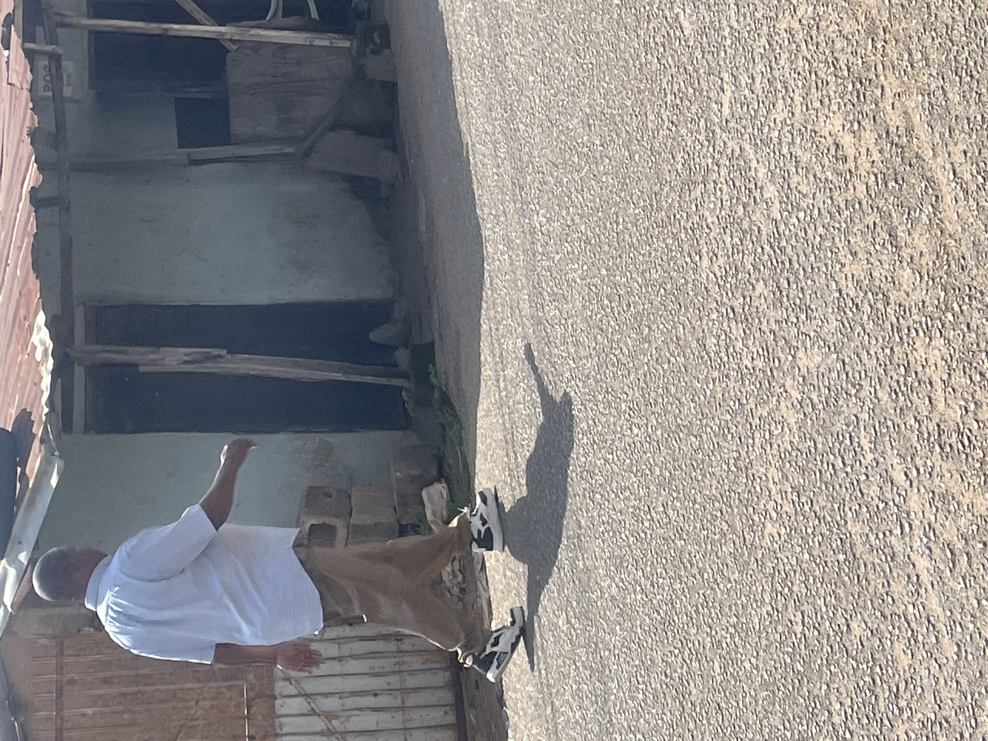 A man in a white short-sleeved collared shirt and tan pants walks away from the camera on a paved surface in front of a light blue building with a corrugated metal roof. The building appears to be made of rough materials, with visible wooden supports and a large open doorway. The man is wearing black and white sneakers and casts a shadow on the ground. The scene is consistent with a roadside environment in a less developed area, potentially on the outskirts of Jos, Nigeria, given the building style and materials.