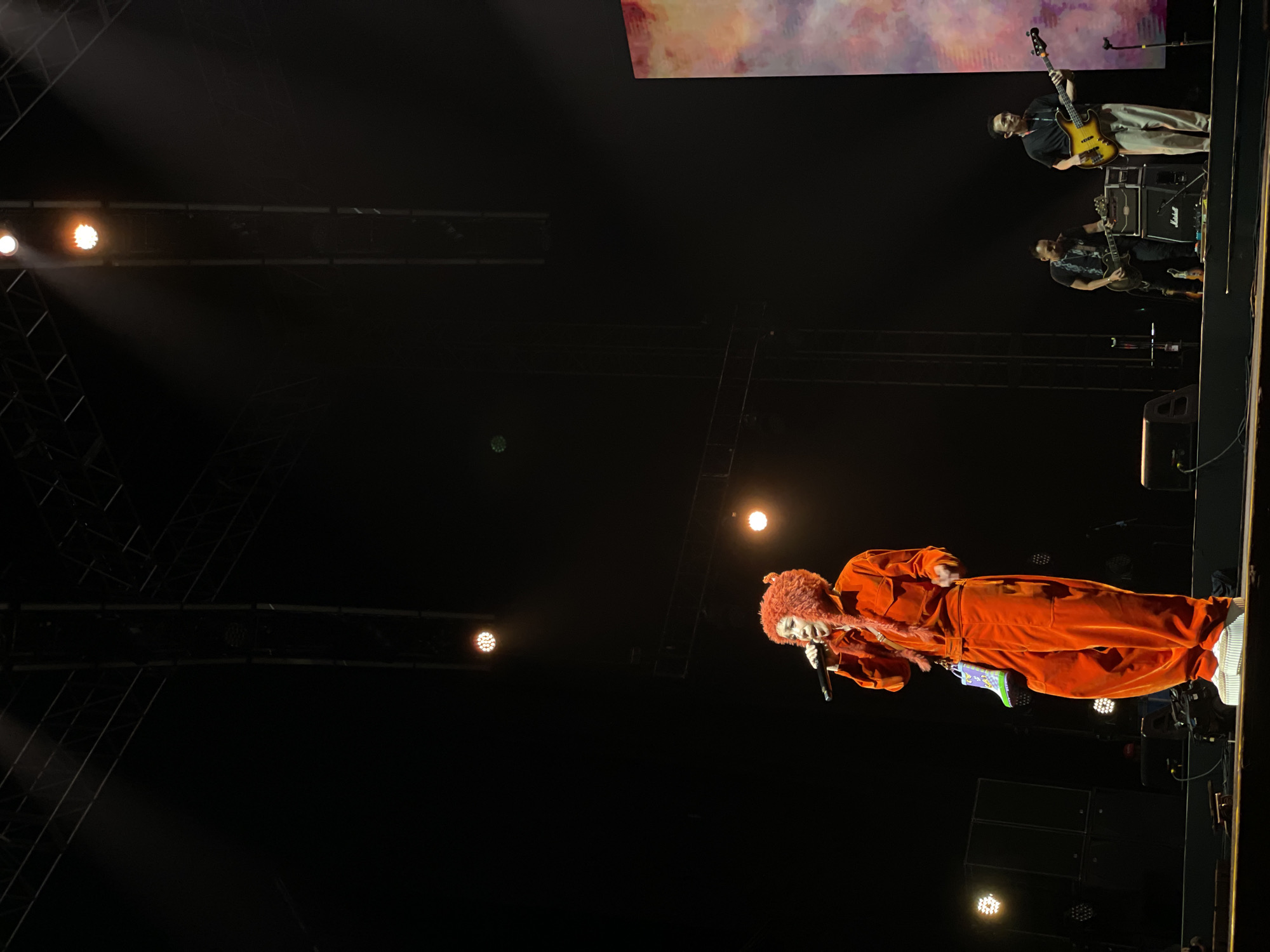 The image depicts a live music performance. A vocalist, wearing an orange jumpsuit and a distinctive furry hat or wig, is singing into a microphone. Behind the vocalist and slightly to the right, two musicians are playing instruments, one with a bass guitar and another with an electric guitar and a Marshall amplifier is visible. The scene appears to be a stage setup with lighting rigs above. A screen is positioned at the top of the frame. The location is Salembaran, Indonesia.