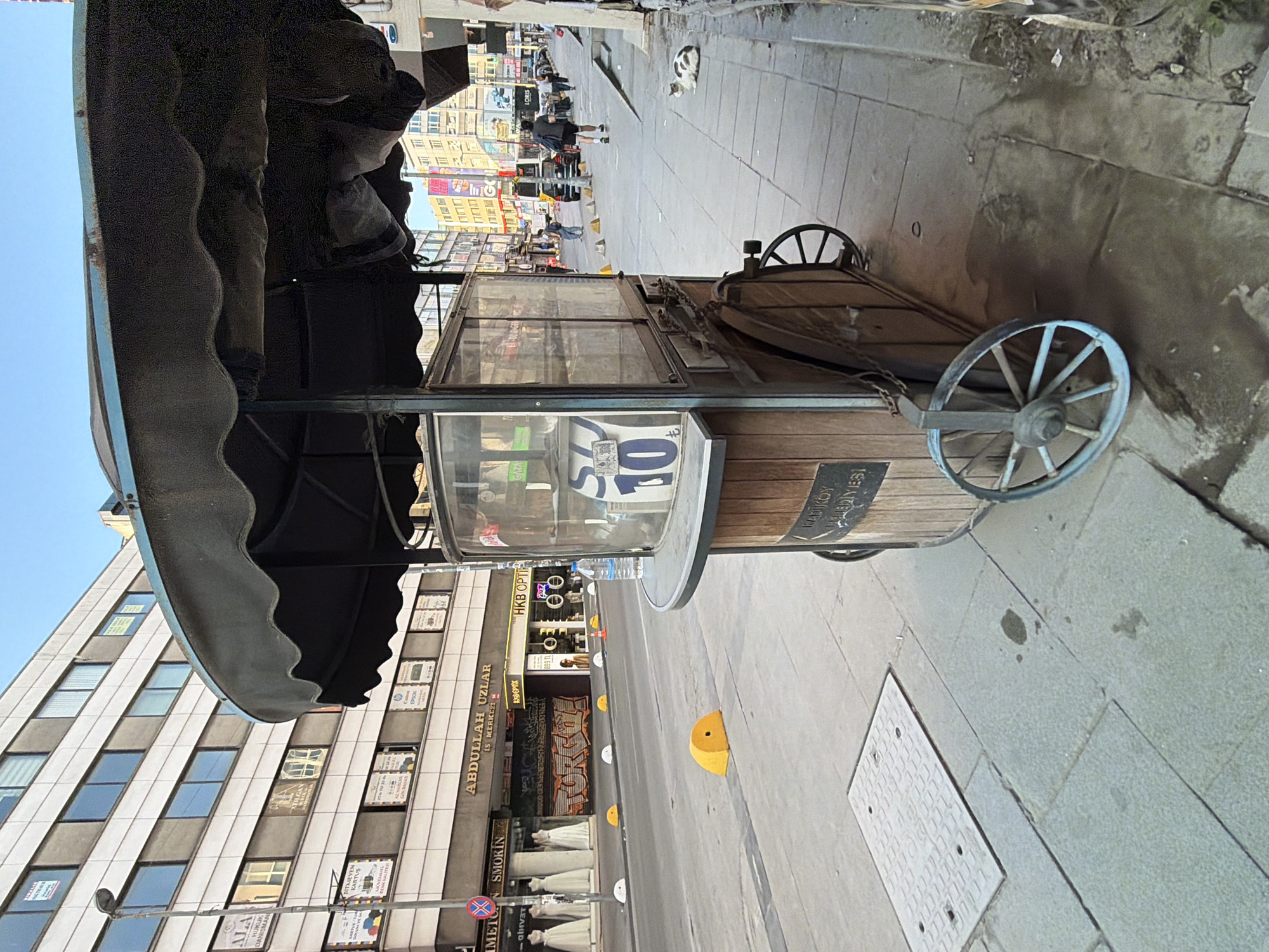 A street scene in Kadıköy, Turkey, features a weathered wooden and metal food cart with a black canopy. The cart displays a sign with the number "10" in blue on a white background. The cart is positioned on a sidewalk adjacent to a street. Across the street, buildings with multiple floors and windows are visible. Yellow, semi-circular traffic barriers are placed along the curb. Further down the street, pedestrians are walking, and vehicles are present. A white and brown cat is resting on the sidewalk. The sky is clear and blue.