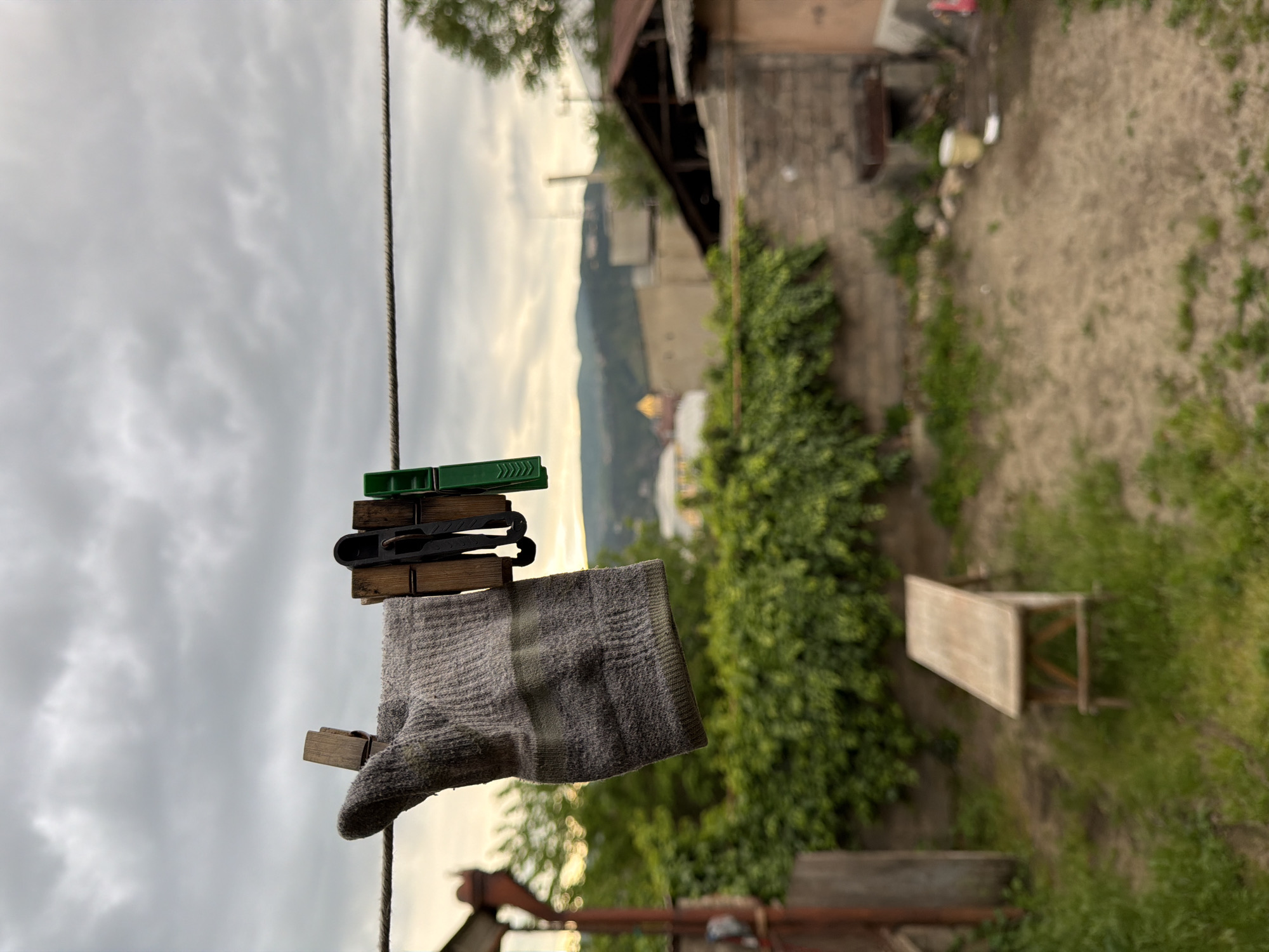 A gray sock is suspended from a clothesline by three clothespins (one green plastic, one black plastic, and one wooden). The background features a cloudy sky, a building with greenery growing on the wall, and a small wooden bench. The location is Tbilisi, Georgia, evidenced by the distinct architecture and landscape. The sock appears damp, suggesting it was recently washed. The background is slightly out of focus.