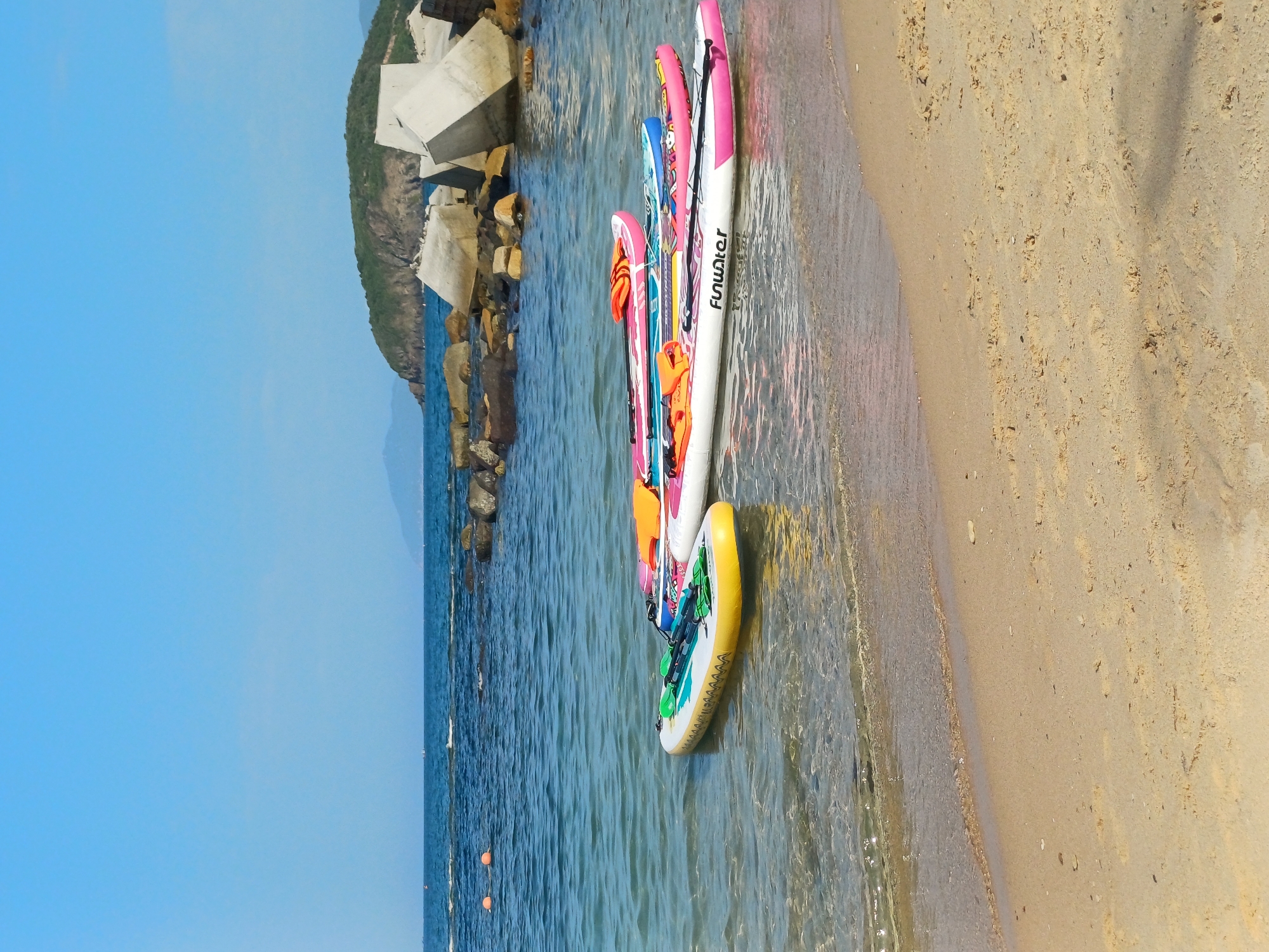 The image depicts a beach scene in Nha Trang, Vietnam. In the foreground, a sandy beach meets clear, shallow ocean water. Four paddleboards are floating on the water near the shore. The paddleboards are of various colors and designs, with some featuring orange life vests. In the background, a rocky breakwater or pier made of large, angular concrete blocks extends into the sea, with a green, tree-covered island or peninsula visible further in the distance. The sky is a clear, bright blue, with a few distant boats on the horizon.