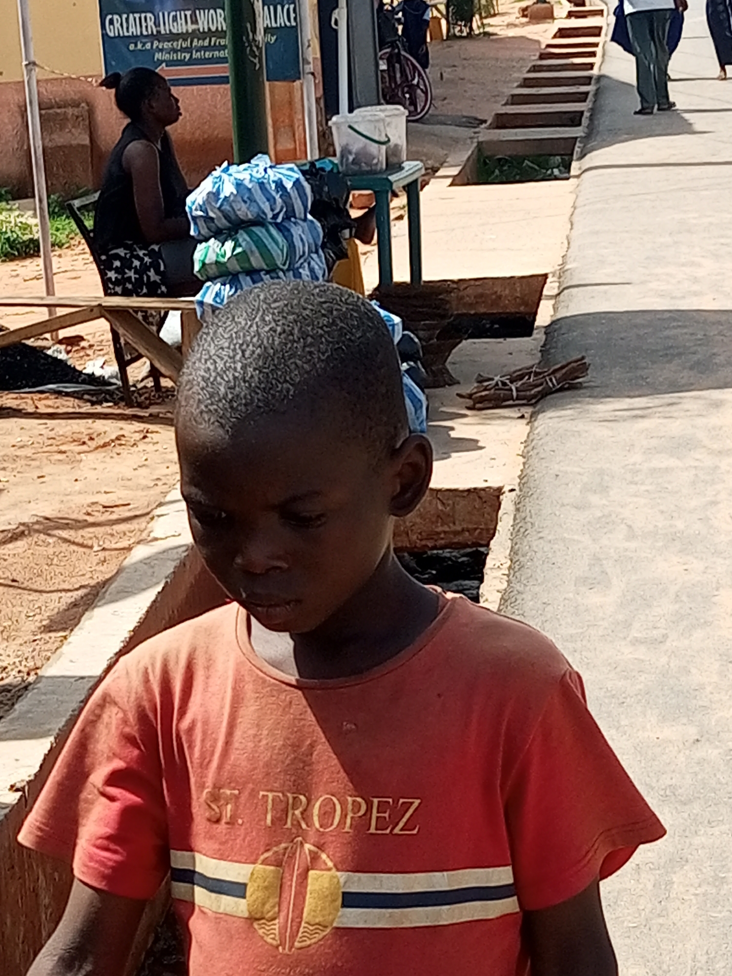 This is a medium-shot, eye-level, outdoor, natural-light photograph taken in the city of Gombe, Nigeria. The primary subject is a young Black boy in the foreground, facing slightly to his right and looking downwards. He is wearing a short-sleeved, orange t-shirt with the words "ST. TROPEZ" printed across the chest in a yellow font, and a circular emblem below it featuring a stylized representation of a surfboard. His hair is short and dark, and his skin tone is dark brown.
To the left of the boy, slightly out of focus, is an adult Black woman sitting on a chair. She is wearing a black sleeveless top and a patterned skirt. Her hair is pulled back in a bun. Behind her is a blue and white striped sign with text that is partially obscured but appears to say "GREATER LIGHT-WOR."
In the middle ground, to the right of the woman, is a stack of blue and white striped bags, possibly merchandise, placed on a wooden structure. Further to the right, a concrete sidewalk or path runs alongside a drainage ditch. A few sticks tied together with twine are visible lying on the edge of the concrete. In the background, a portion of a bicycle and a green table with a white container on it are visible. The ground appears to be dirt or sand. The overall impression is a scene from a street market or roadside area in Gombe, Nigeria.