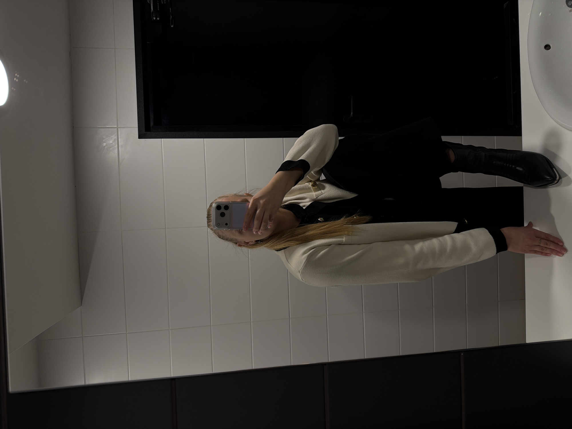 A person is taking a selfie in what appears to be a bathroom. The person is wearing a beige and black jacket, black pants, and black boots. The person is holding a phone in front of their face, obscuring their features. The background consists of white tiled walls and a black cabinet. A white sink is visible on the right side of the frame. The location is undetermined, and there is no visible information indicating it is in Izhevsk, Russia.
