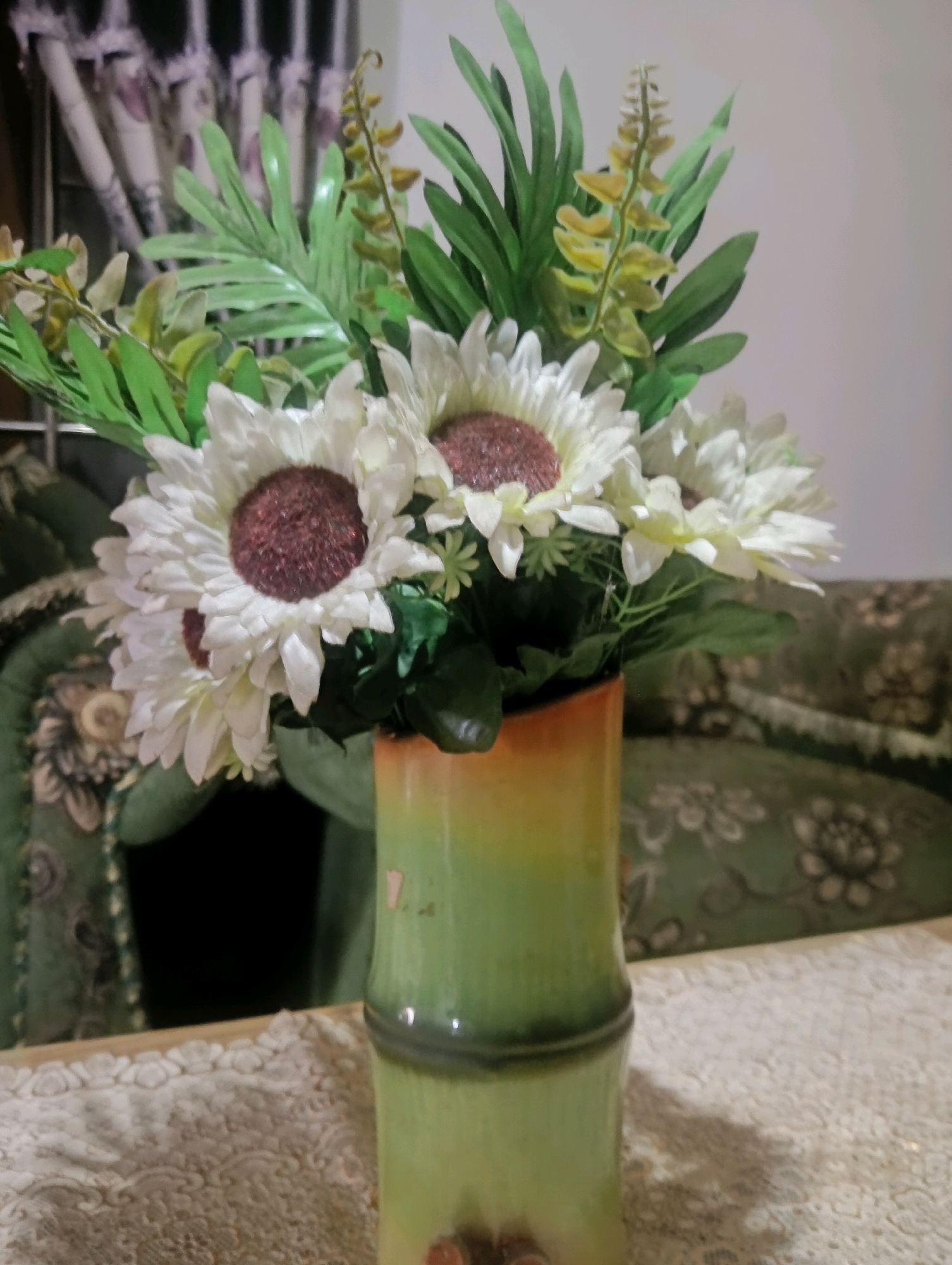 A close-up, eye-level shot captures a vase of artificial sunflowers on a table. The vase is green and orange, with a bamboo-like texture and segmentation. The sunflowers are white with dark brown centers and are arranged with various green artificial leaves and small yellow sprigs. The vase is placed on a textured, off-white tablecloth with a floral pattern. In the background, a dark sofa with a green floral pattern is visible. The overall lighting is dim. The image is taken in Kromengan, Indonesia.