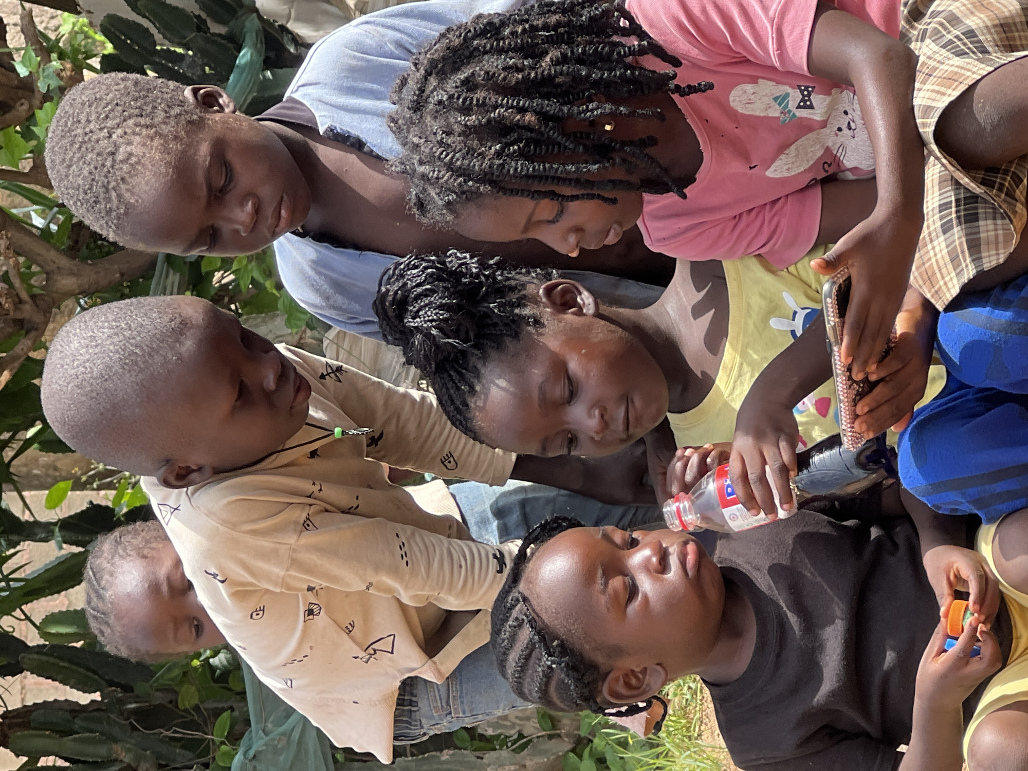 The image captures a group of five children outdoors, appearing to be in a rural or semi-urban setting that could be the city of Jos, Nigeria. The children, all of African descent, are of varying ages and are clustered together, engaging with a smartphone.
In the foreground, a young child wearing a black t-shirt and yellow shorts sits on the ground, holding two small, colorful blocks. This child is looking down at the smartphone. To the right, another child, wearing a pink shirt with a rabbit graphic, is also looking at the smartphone, with their arm resting on their lap. This child has braided hair. Between these two children, a third child in a yellow shirt is holding a bottle of water and is also looking at the phone. This child has their hair in braids.
Behind the foreground children, two older children are visible. One, wearing a light-colored patterned shirt and jeans, is bending over and looking towards the smartphone. The other, wearing a blue shirt, is standing behind the first older child, also looking towards the phone. A hint of a cactus and leafy green vegetation forms the background, suggesting an outdoor environment. The lighting indicates it is daytime.