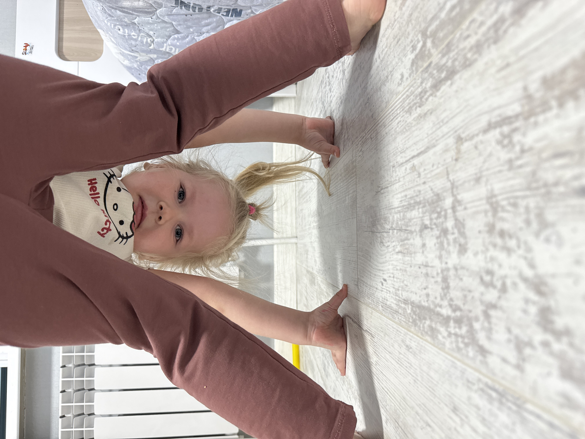 A young child with blonde hair in a ponytail is positioned upside down, her hands placed on a light-colored wooden floor. She is wearing a white shirt with a cartoon character and mauve-colored pants. The child's face is visible, with blue eyes looking directly at the viewer. The background includes a white crib and a mattress with the word "Neptune" partially visible. The location is likely indoors, possibly in a home or nursery. No specific details directly imply the city of Gornyi (Dvurechenskoe S/P), Russia, are apparent.