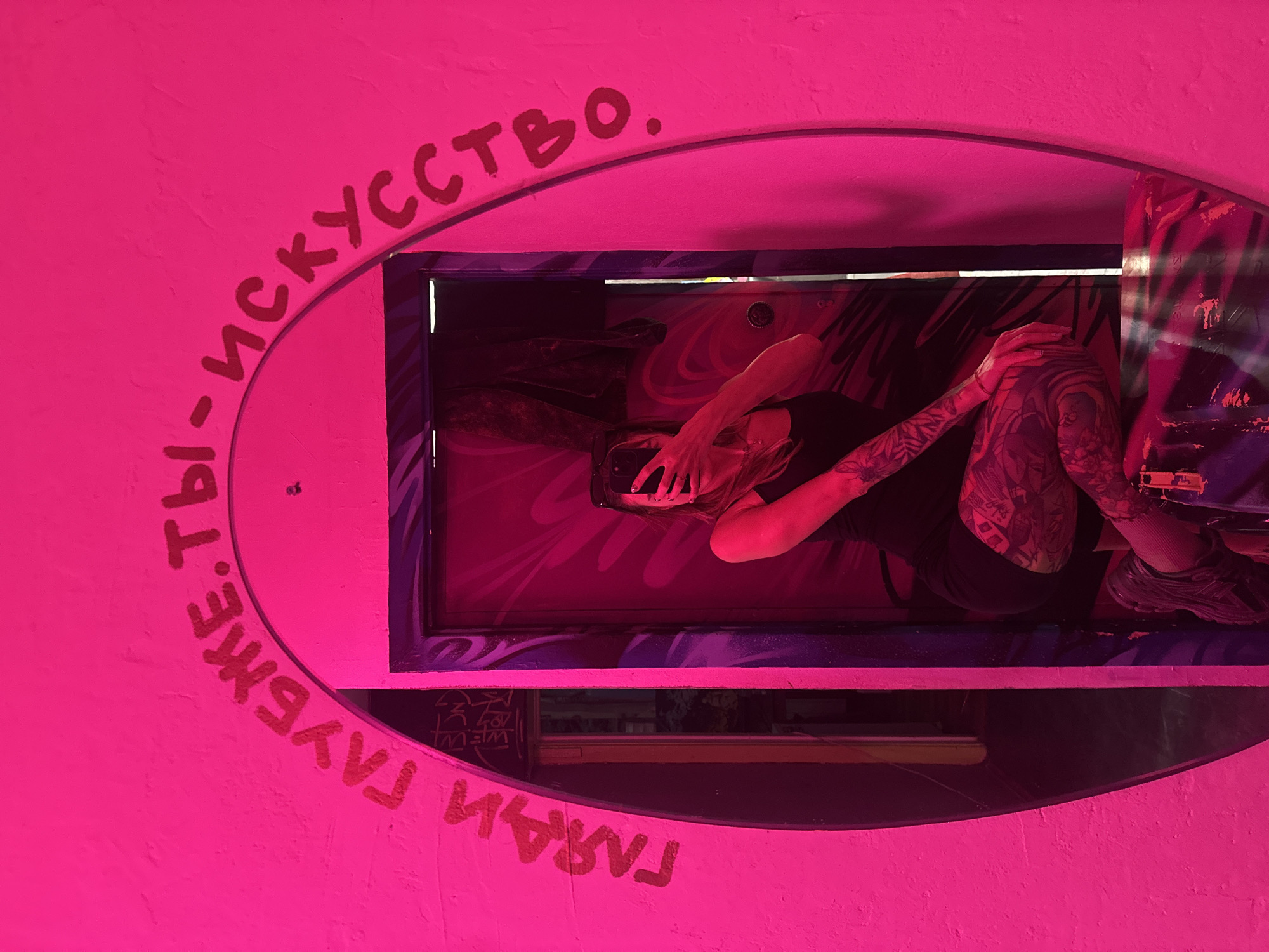 The image depicts a person reflected in an oval mirror inside a structure painted bright pink, likely in Moscow, Russia. The person in the reflection is holding a cell phone and covering their face with their hands. They are wearing a black shirt, pants, and shoes, and their arms and legs are heavily tattooed. The wall surrounding the mirror is painted pink with text written in Russian above and below the mirror opening. Inside the mirror and above the frame, there are some more colors and a red wall. The whole scene is inside a building or structure that has been decorated with vibrant and bold colors.
