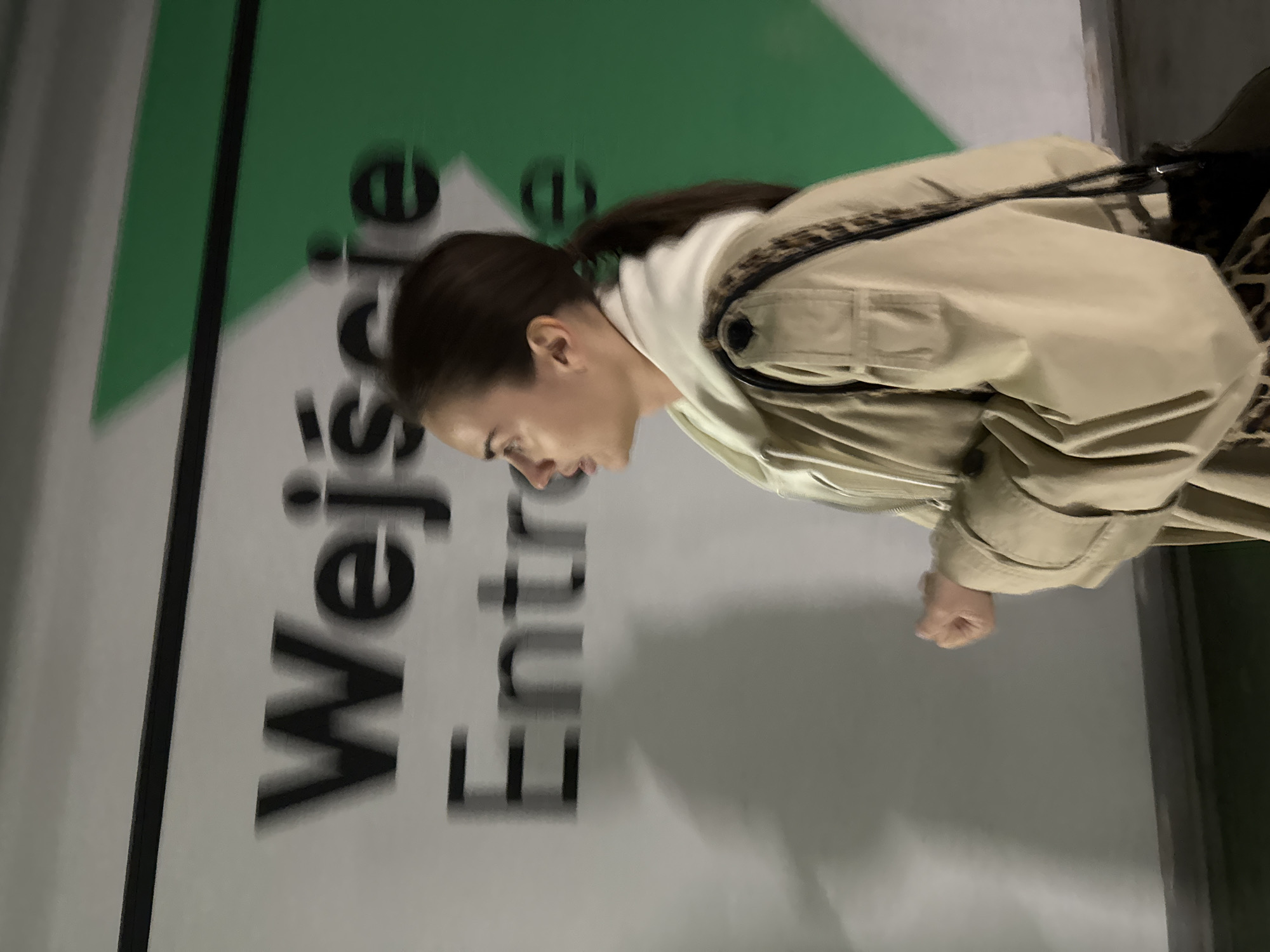 An indoor, medium shot captured from a slightly high angle shows a woman in profile, looking downwards. She is positioned towards the right side of the frame, with her head and upper torso visible. Her dark hair is pulled back into a ponytail. She wears a light beige or khaki trench coat over a white hooded top. A strap with a leopard print pattern is visible across her right shoulder, suggesting a bag. Her right hand is partially visible, clenched into a fist at waist level.
The background consists of a light gray wall featuring a green geometric design, possibly a large triangle or arrow. Overlaid on this wall are large, bold black letters. The text "Wejście" (Polish for "Entrance") is prominently displayed, followed by "Entre," which is a common term for "Enter" or "Entrance." The presence of "Wejście" strongly indicates the location is in Poland, specifically Warsaw as per the provided context.
The overall image has a slight motion blur, particularly noticeable in the text, suggesting either subject movement, camera movement, or a longer exposure in low light conditions. The lighting appears artificial, contributing to the slightly muted color palette. The woman's expression is neutral and focused, perhaps pensive, as she looks down. The scene appears to be an entrance area in a public building or event space.
