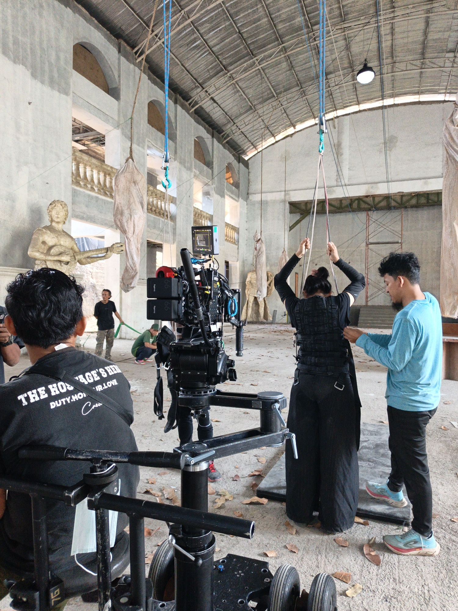 The image depicts a film set in an abandoned industrial building, possibly in Cibogo, Indonesia, with a concrete floor littered with fallen leaves. In the foreground, a large professional camera rig is set up on a dolly. A person in a black t-shirt with white text reading "THE HOSTAGE'S HERO" is positioned behind the camera, seemingly as part of the crew.
In the mid-ground, a woman wearing a black vest and dark pants is being assisted by a man in a blue long-sleeved shirt. The woman's arms are raised, and she appears to be tethered by ropes hanging from the ceiling. Several other figures are visible in the background, including a person in a black t-shirt and camouflage pants holding a green strap, and another person crouching.
The building's architecture features large arched openings and a high, corrugated metal roof supported by exposed beams. Statues and hanging fabric objects are also present within the space, suggesting a deliberate artistic or thematic arrangement for the filming. The overall scene conveys an organized, yet potentially dramatic, production environment.