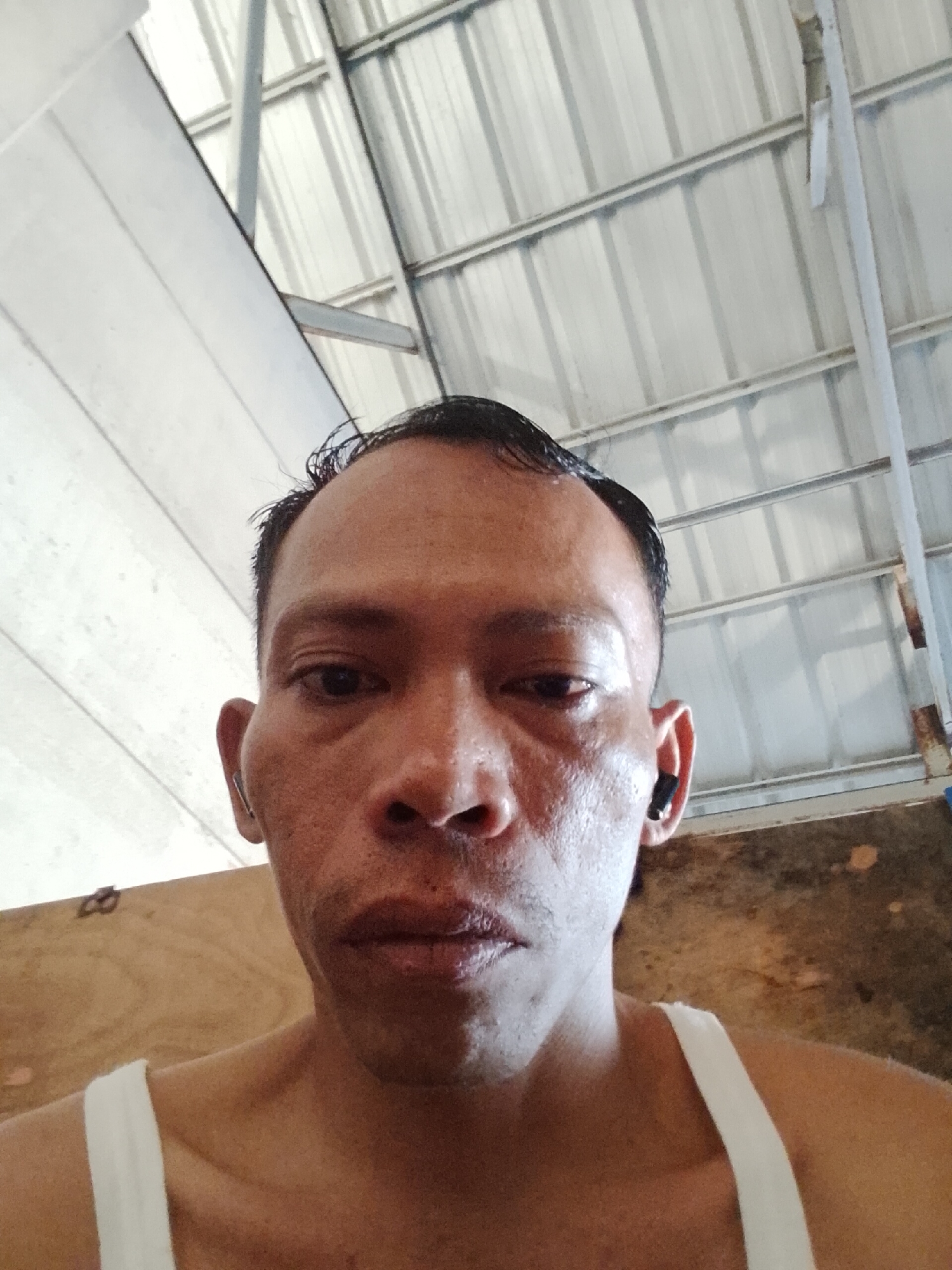 A close-up, low-angle selfie is captured from the chest up, featuring a man with dark, short, wet hair. He is wearing a white tank top and has a dark earbud in his right ear. The background is an indoor setting with a corrugated metal ceiling and what appears to be a wooden wall or partition. The lighting suggests an overcast day or an indoor space with indirect light. The man's face is the primary focus, with his expression neutral. The overall scene is informal and personal. The image was taken in Bukit Ketok, Indonesia.