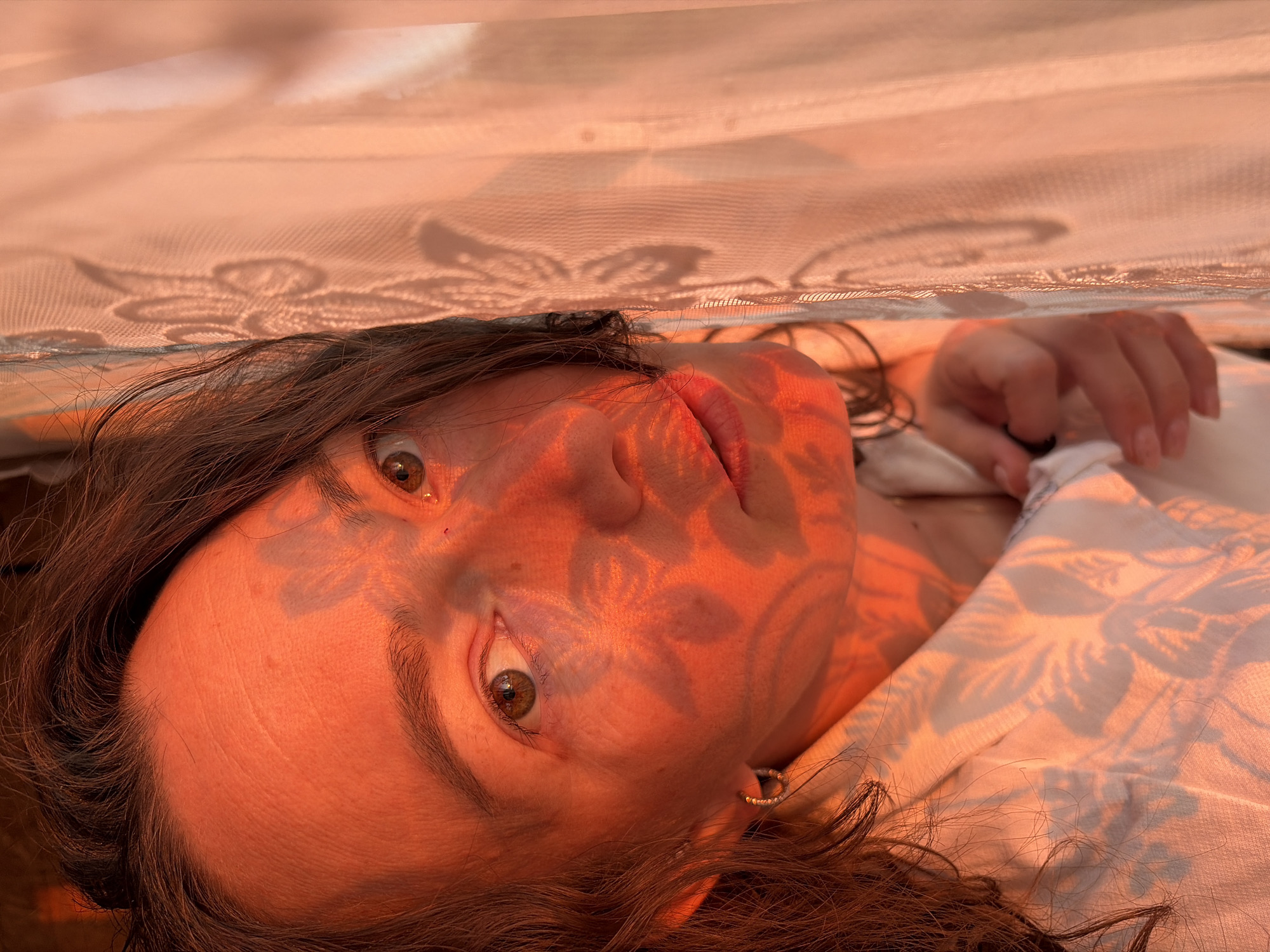 A close-up, rotated photograph captures a person's face and upper body, illuminated by the warm, golden-orange light of either sunrise or sunset. The individual, appearing to be female with dark brown hair, is lying down and looking directly at the viewer with wide, focused brown eyes, conveying an expression of surprise or direct engagement.
Intricate floral patterns, created by shadows cast from a translucent, lace-like fabric draped above, fall across her face, hair, and the light-colored, floral-patterned top she is wearing. Her lips are slightly parted, and a small, beaded hoop earring is visible on her right ear. Her right hand is partially visible in the upper right, resting on the fabric. The light source appears to be direct and strong, highlighting the textures of her skin, hair, and the fabric, and contributing to the dominant warm color palette of oranges, reds, and browns. The setting, possibly indoors or under a canopy, creates an intimate and contemplative mood. The location context is Ольховка, Russia. There is no visible text.