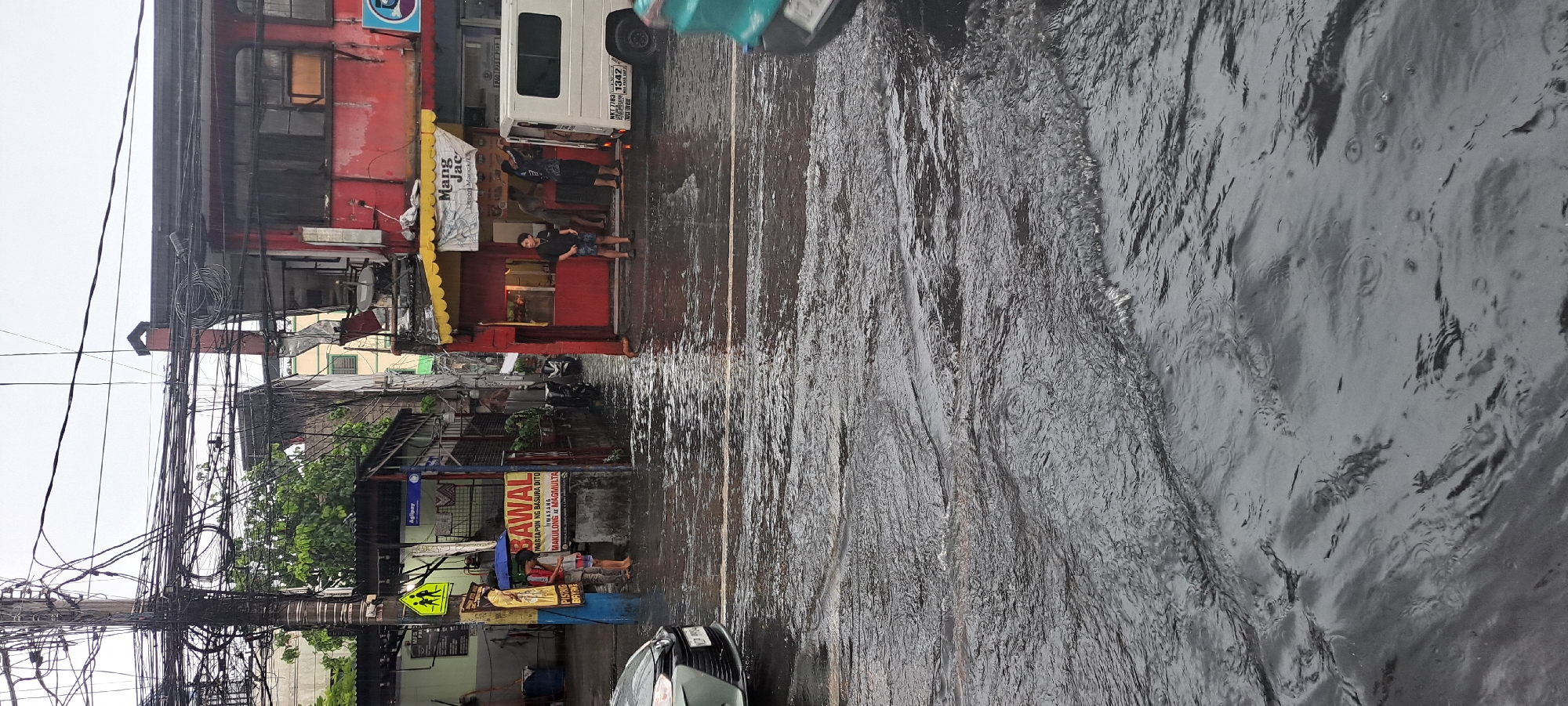 A flood scene is depicted in the city of Mandaluyong, Philippines, with water covering the street and sidewalk. The sky is overcast, suggesting recent or ongoing rain. On the left side of the street, a sign with the word "BAWAL" (meaning "prohibited" in Tagalog) is visible, along with text indicating that littering is prohibited and subject to fines. Two individuals are standing under a blue umbrella, facing the sign. Further down the street, on the right side, a red building with a yellow awning displays the name "Mang Jac." Several people are standing in front of this establishment, and a white van is parked nearby. A light green car is in the foreground, partially submerged in the floodwater. The street is filled with rippling water, indicating movement, possibly from vehicles or the rain. The environment appears to be an urban setting with electrical wires strung overhead.