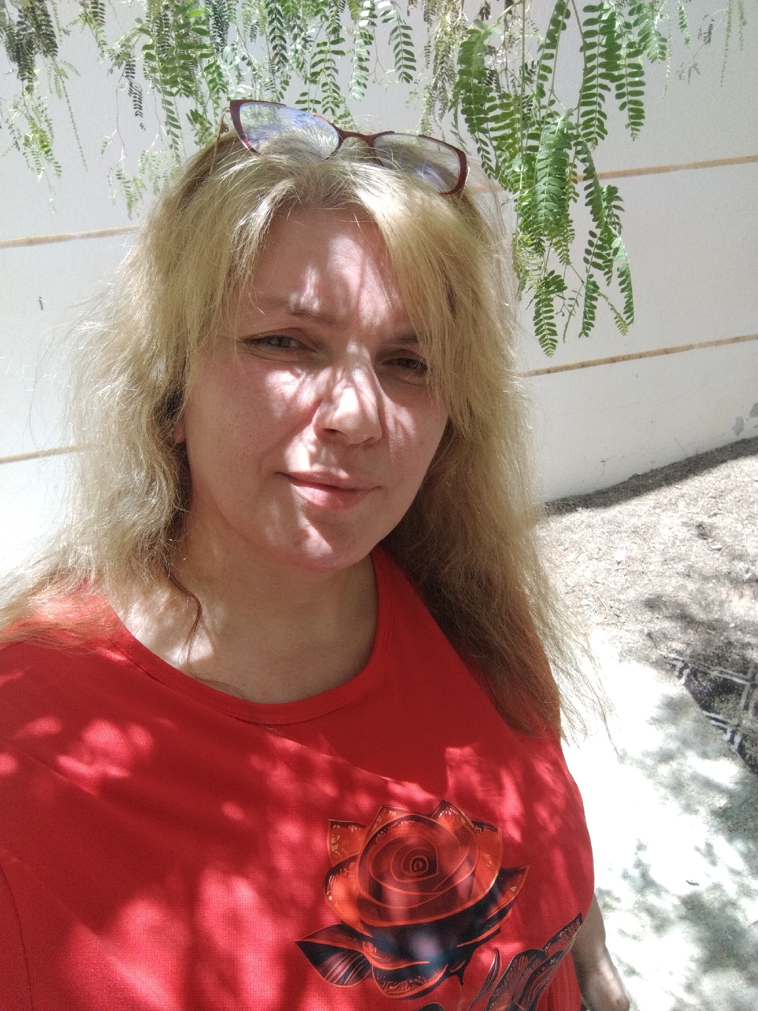 The image depicts a woman in a red shirt with a rose design, standing outdoors in dappled sunlight. She has fair skin and blonde hair, with glasses perched on top of her head. The background features a white wall with hanging green plants. The ground is covered in light-colored gravel or dirt. The photograph was taken in Al Mas'oudi, United Arab Emirates.