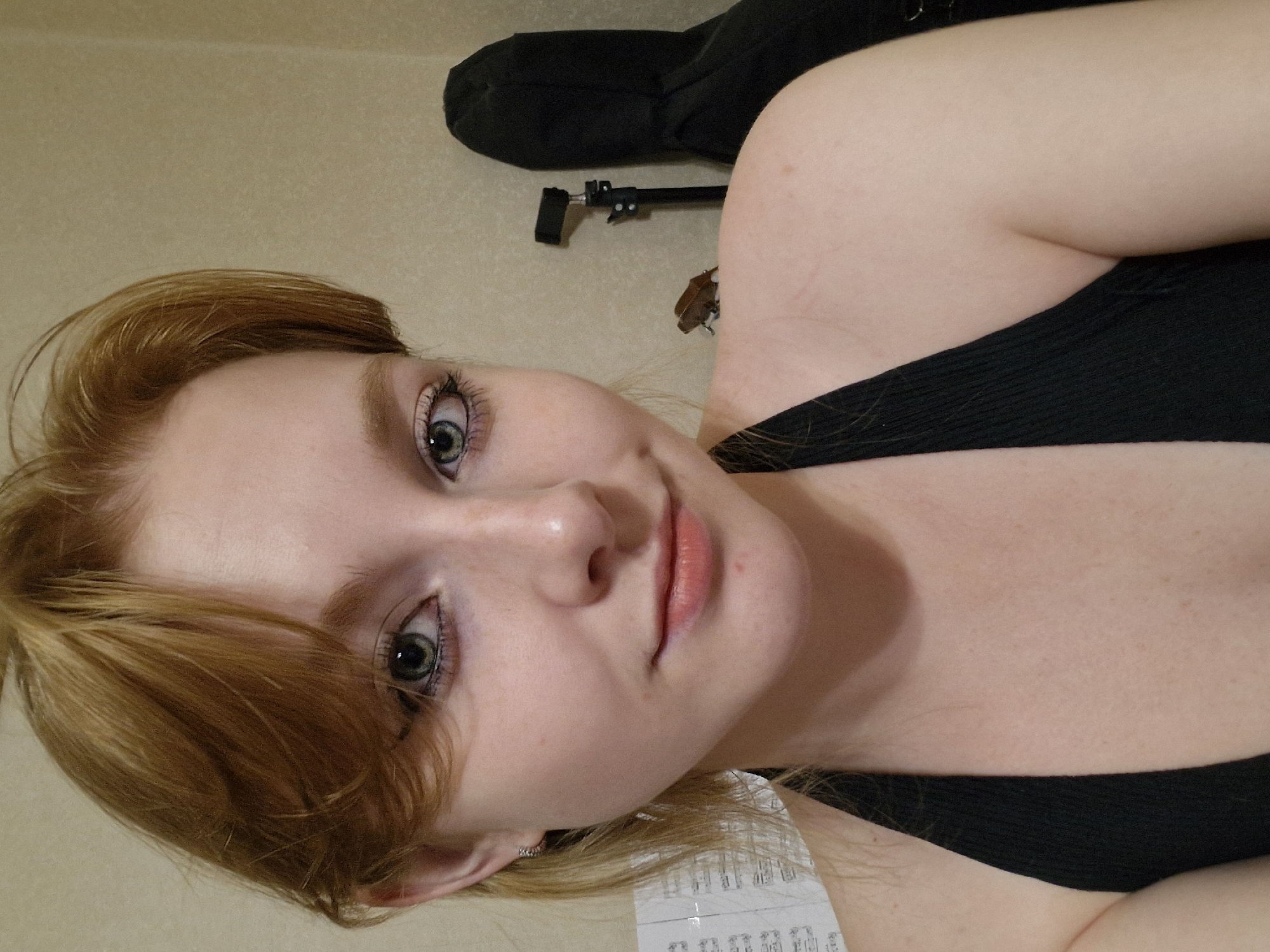 The image is a close-up selfie of a young Caucasian woman with auburn hair, fair skin, and green eyes. She is wearing a black halter top. Her makeup includes eyeliner and mascara. In the background, to the right, is a black case, possibly for a musical instrument, and a black tripod with a small light attached. On her right shoulder, a small brown amphibian, likely a frog or toad, is perched. The wall behind her is a textured beige. A sheet of music is partially visible on the left side of the frame. The image does not contain information that would allow for identification of its location within Yekaterinburg, Russia, or any specific city.