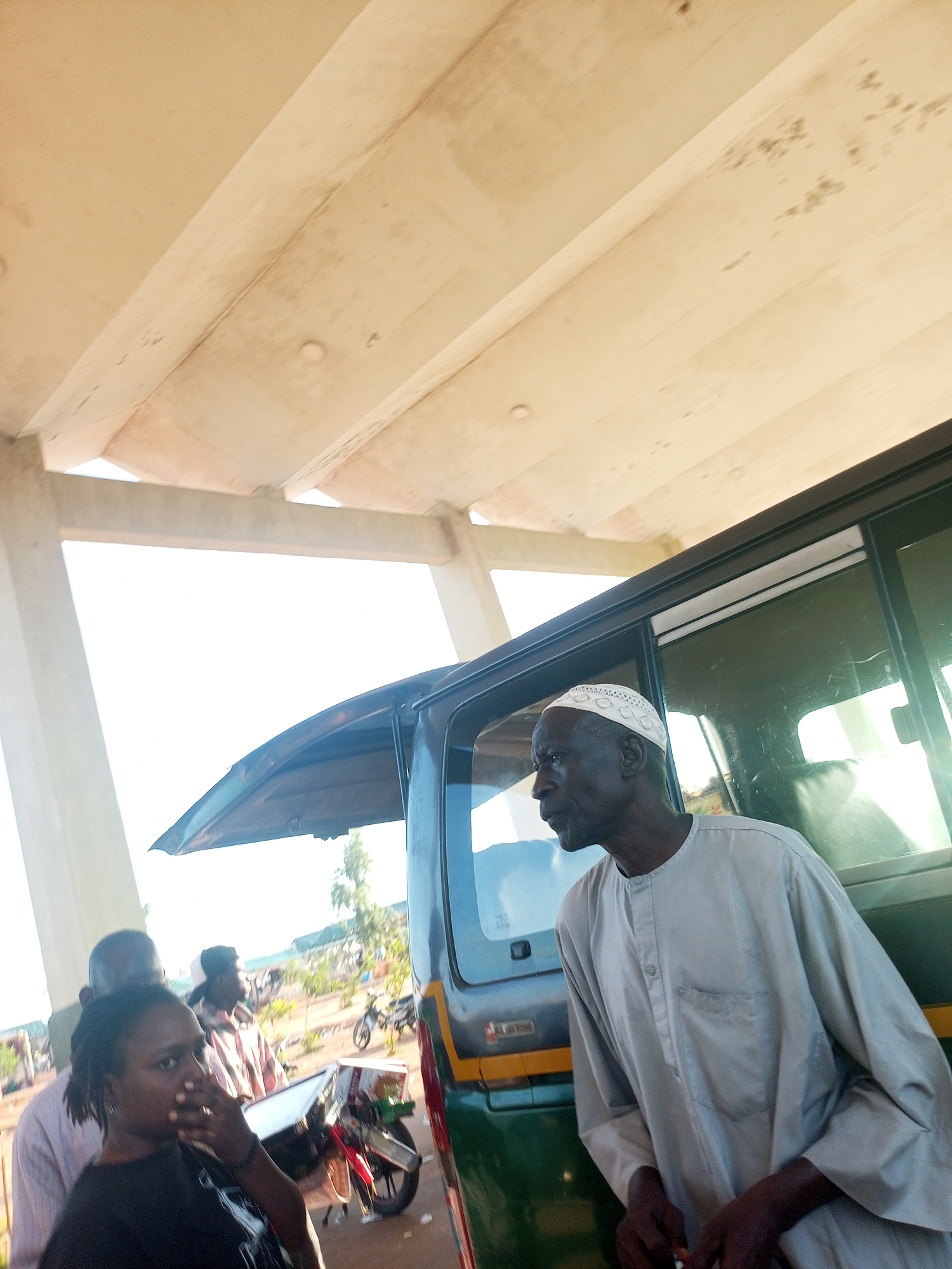 An outdoor scene under a concrete structure reveals an older man in a traditional white cap and light grey tunic standing next to a green vehicle. Another person, a woman in a black top, is partially visible in the foreground, along with a person in a checkered shirt. Behind them, a motorbike is parked. In the background, hints of a cityscape suggest an urban setting. The specific city and region are unknown.