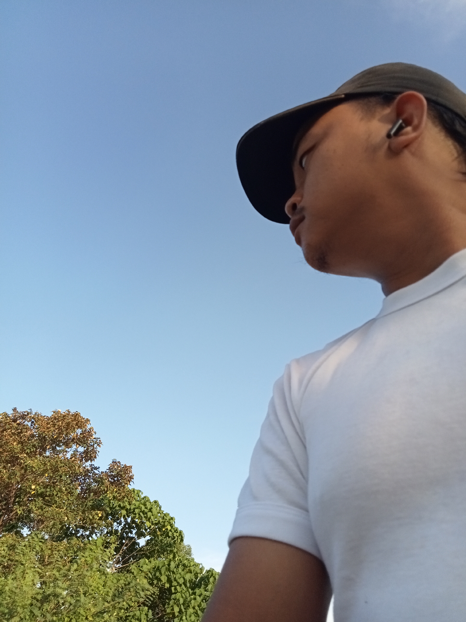 The image shows a low-angle outdoor shot of a person wearing a white polo shirt and a black baseball cap, looking to their right. The person has an earbud in their right ear and appears to have a small amount of stubble on their chin. Behind them, the sky is a clear, light blue. In the lower left of the frame, the tops of green and brown trees are visible. The scene suggests an open, possibly rural, setting. The provided location of Teluk Kenidai, Indonesia, aligns with the natural elements seen in the image.