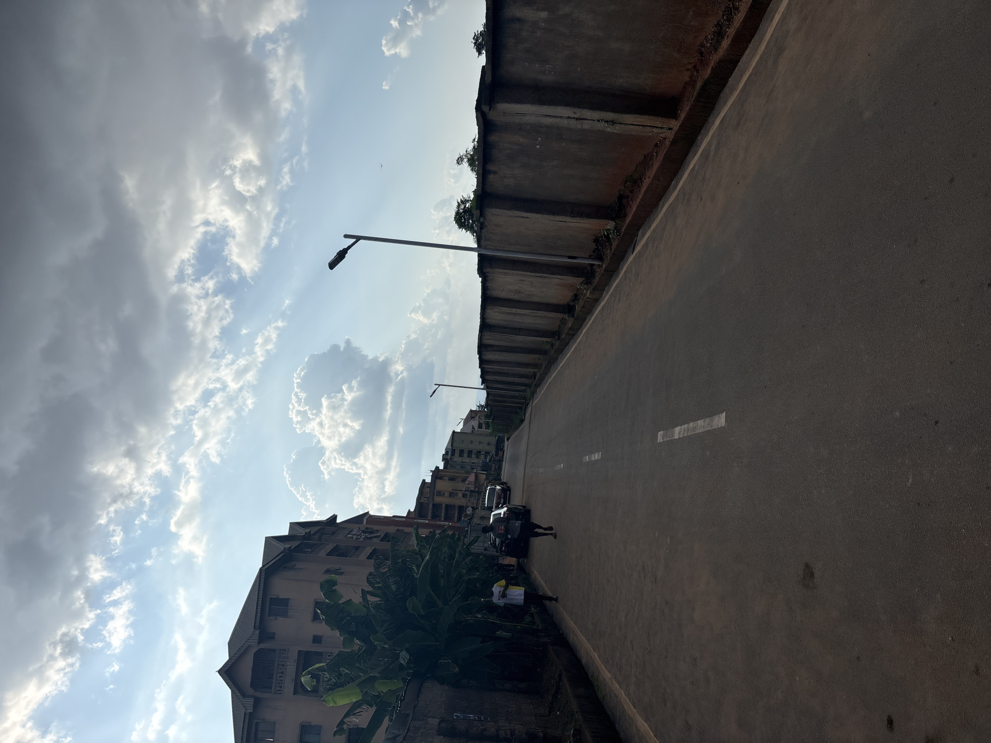 This is a full shot of a paved road in the city of Onitsha, Nigeria. Two men are jogging down the road. The man on the left wears a white t-shirt with yellow trim and the man on the right wears a black t-shirt and black shorts. The road has a white stripe down the middle and a lighter stripe on the right side. On the left side of the road is a tall, multi-story building and a wall with a large banana tree. On the right side of the road is a tall, concrete wall with trees visible above it and a street light. The sky is partly cloudy with sunlight breaking through.