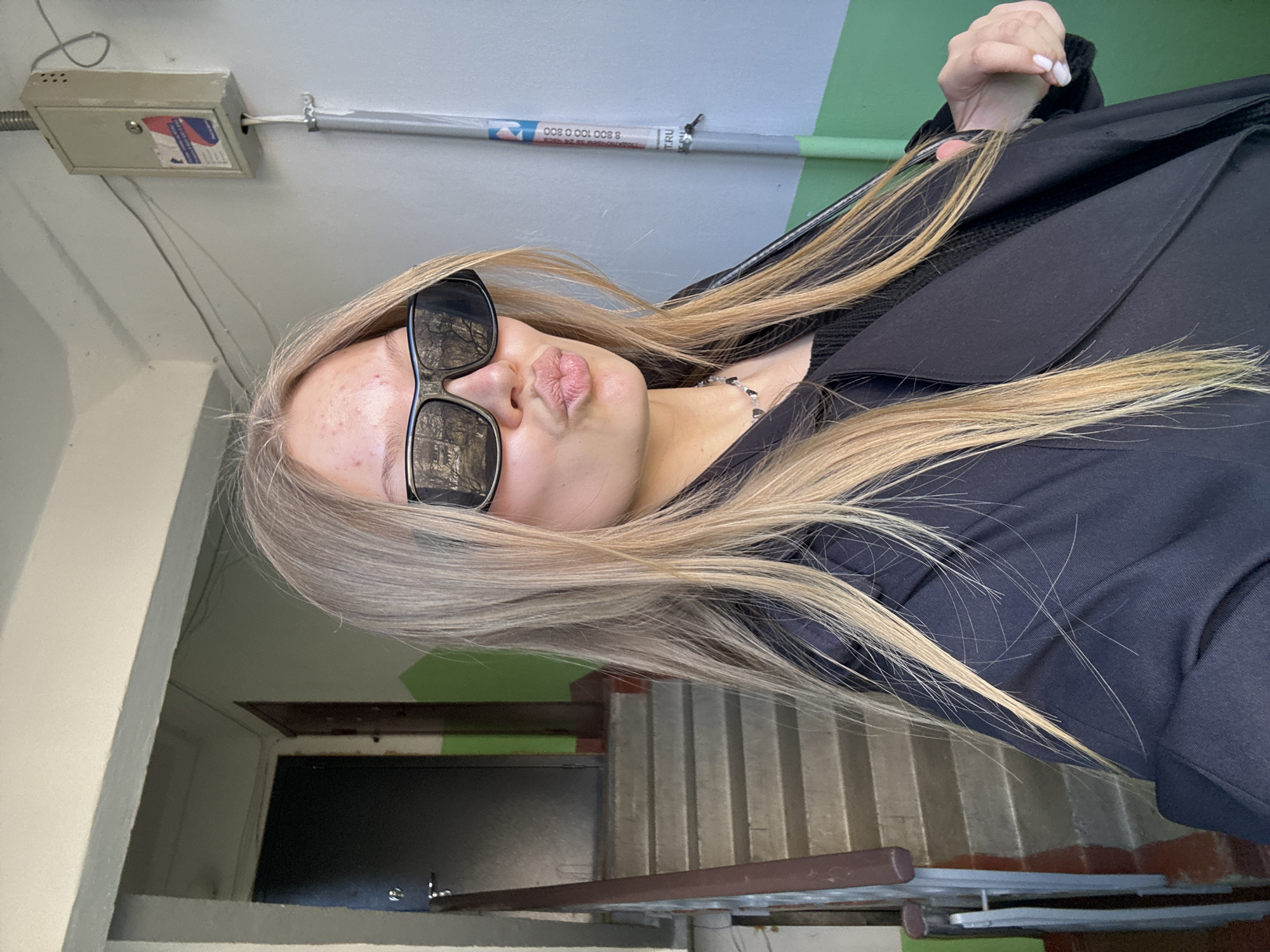 A photograph taken in a stairwell-like location in Chaikovsky, Russia. The subject is a blonde woman wearing dark sunglasses and a black coat. She has her hair in a cascade down her side. She is pursing her lips in a kissing gesture. The background includes stairs and a door, with a green and white wall overhead. Above her are metal pipes running along the ceiling. An electrical box is mounted above.