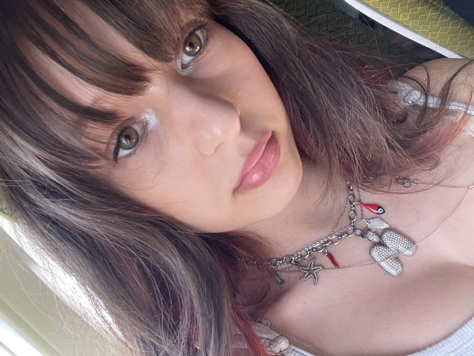 A close-up selfie captures a young female with long, brown hair featuring subtle reddish-pink highlights and blunt bangs. Her hazel/green-brown eyes are open and directed towards the camera, while her lips are full and glossy pink. She wears a light-colored, possibly white, top with a visible strap on her left shoulder. Adorning her neck are multiple necklaces: a prominent silver-toned chain adorned with various charms, including a large pave-set initial "M", a textured starfish, a red chili pepper (cornicello), and a red fish charm detailed with an evil eye motif. A thinner, delicate gold-toned chain is also visible beneath the main necklace. The background is softly blurred, revealing a bright light source on the upper left and a hint of a greenish-yellow patterned textile or surface on the right. The scene is located in the city of Tzafria, Israel.