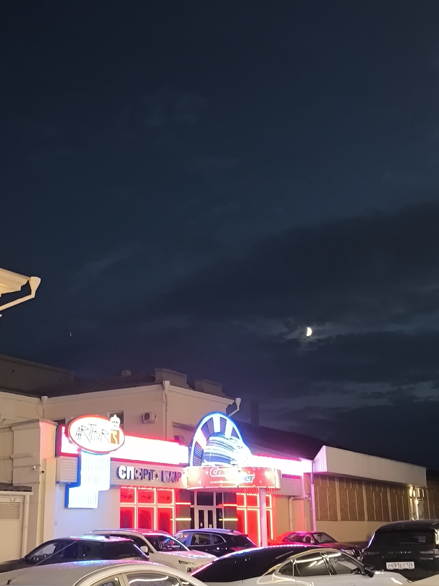 An exterior, vertical, low-angle shot captures a building with bright neon signage at night in Yessentuki, Russia. The building features a prominent sign reading "ARTHUR'S" in red and blue neon lights, with a crown graphic above. Next to it, another sign in Cyrillic characters reads "СПОРТ БАР" (SPORT BAR), also illuminated by neon. The building's facade is white, with red neon lights framing an entrance area. Several cars are parked in front of the building. The sky is dark blue with some clouds, and a crescent moon is visible. A small, white vertical streak, possibly a plane or satellite trail, is also present in the upper left portion of the sky. The overall impression is that of a lively establishment at night.