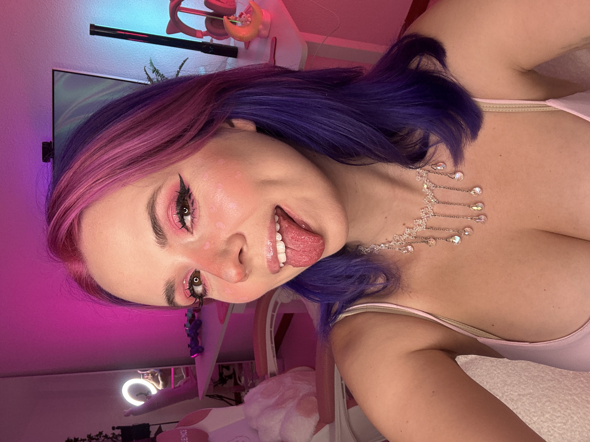 The image is a selfie of a young woman with pink and purple dyed hair. She is wearing a pink top and a delicate necklace with clear teardrop-shaped pendants. She is sticking her tongue out and has pink eyeshadow and blush on her face. In the background, there is a pink gaming chair with a fluffy white pillow, a desk with some electronics, and a monitor. The room is lit with pink and blue neon lights, creating a vibrant and playful atmosphere. The overall scene suggests a modern, stylized, and personal space, possibly a home gaming setup. The image does not provide any specific information or visual cues that would indicate a location within the city of Oviedo, Spain.