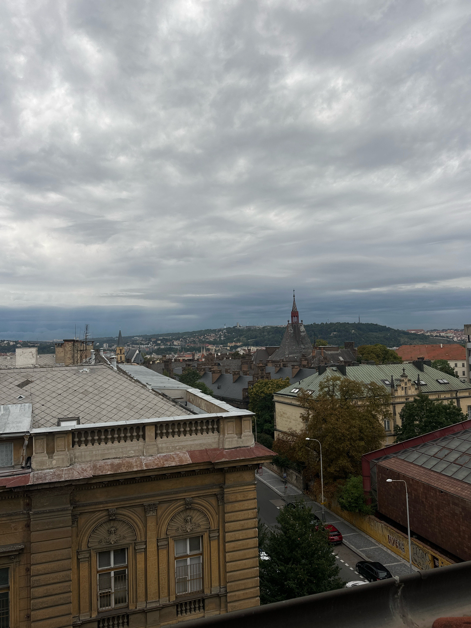 A high-angle, medium shot captures a cloudy sky above a cityscape with an overcast sky. The foreground is dominated by the top of a building with a balustrade and large, ornate windows. To the left, a grey tiled roof extends across the frame. The middle ground features a cluster of buildings with dark, pointed roofs, and a building with a red steeple. A tree with autumn foliage is visible. Further in the distance, a hazy, green hill rises, dotted with buildings and a slender tower. A paved street with parked cars, including a red and a black vehicle, winds through the scene. The overall impression is of an urban environment under a heavy, grey sky.