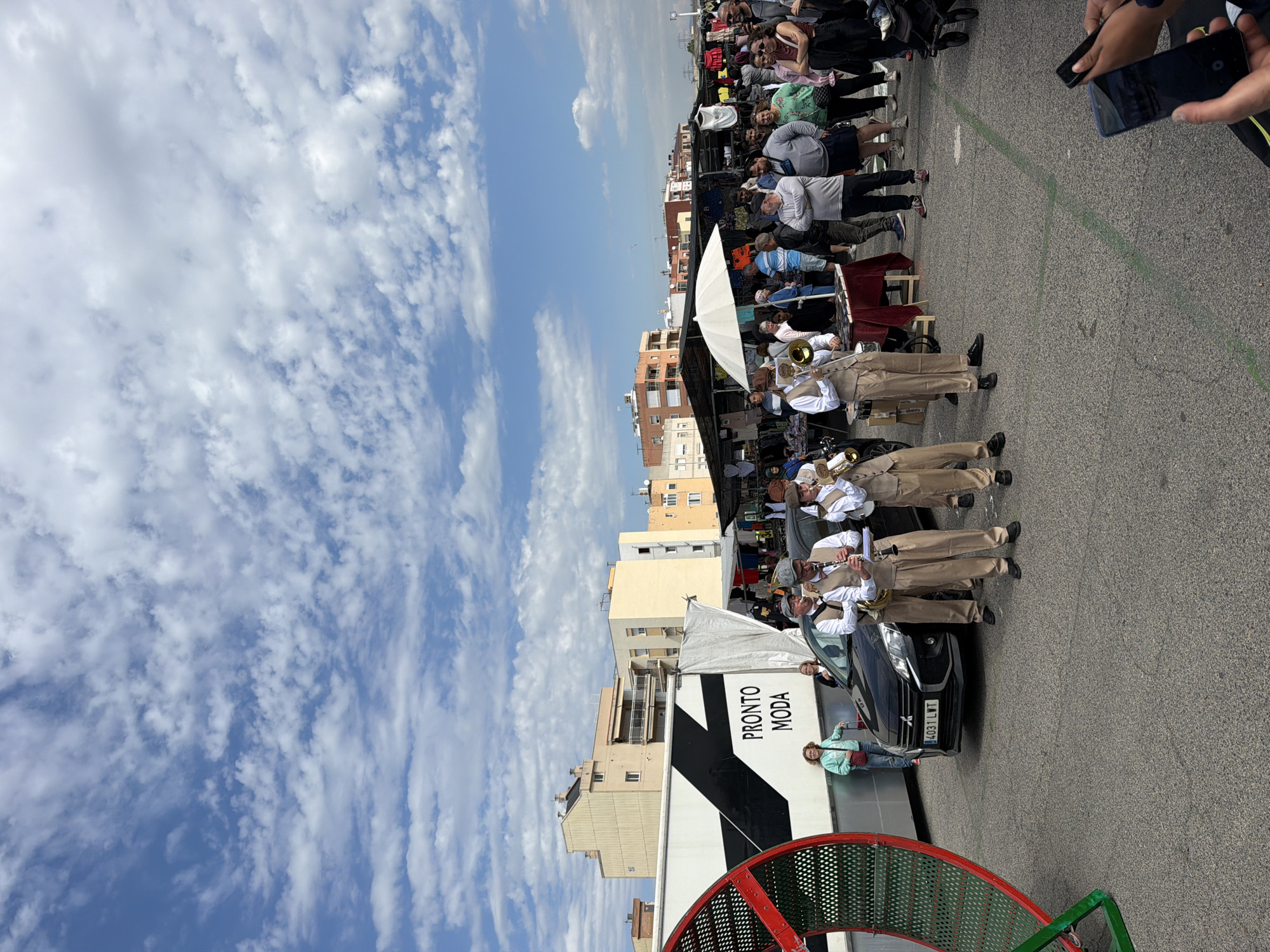 The image shows a street scene in La Canonja, Spain. The sky is partly cloudy. A brass band, dressed in khaki pants, white shirts, and vests, is playing in front of a black car (license plate 4031 LWT) with a PRONTO MODA truck parked behind it. A woman in a light blue jacket stands near the car. A crowd of people are gathered behind the band and stalls can be seen in the background. The edge of a red and green playground apparatus is visible in the foreground. Buildings are seen in the background. A cell phone is visible in the upper right-hand corner of the image.