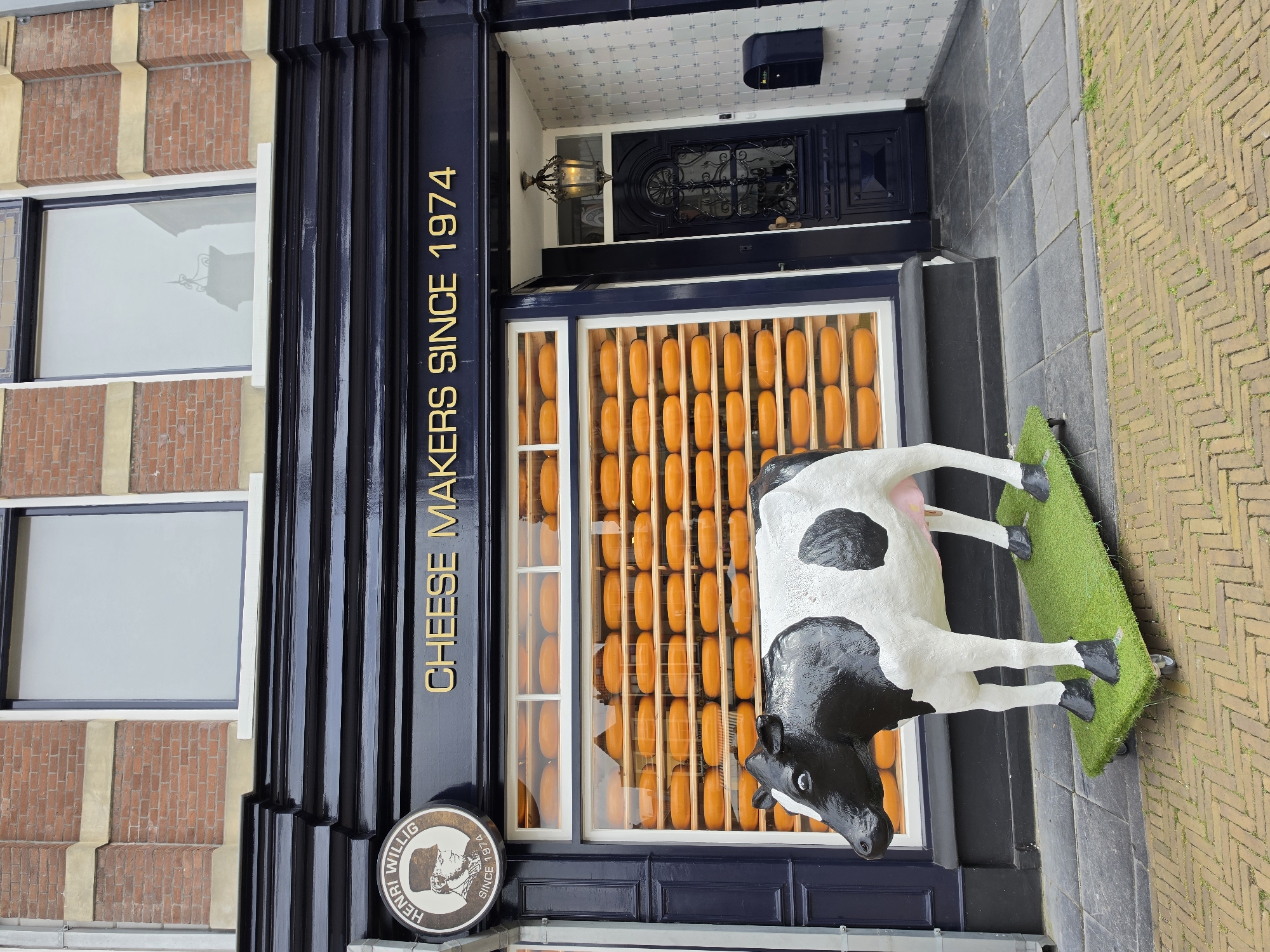 The image shows the exterior of a cheese shop, Henri Willig Cheese Makers Since 1974, located in the city of Delft, Netherlands. In front of the shop, a life-sized statue of a black and white cow stands on a platform covered with artificial grass. The shop has a large display window filled with numerous round, orange cheeses stacked on wooden shelves. Above the window, the shop's name and establishment date are prominently displayed in gold lettering. To the left of the display window, a circular sign with a portrait of a man and the name "Henri Willig" is visible. The shop's entrance is to the right, featuring a dark blue door with intricate metalwork and a vintage-style lantern hanging above it. The facade of the building is adorned with red brick and white trim, and the street in front is paved with cobblestones. A dark blue mailbox is mounted on the wall to the right of the entrance.