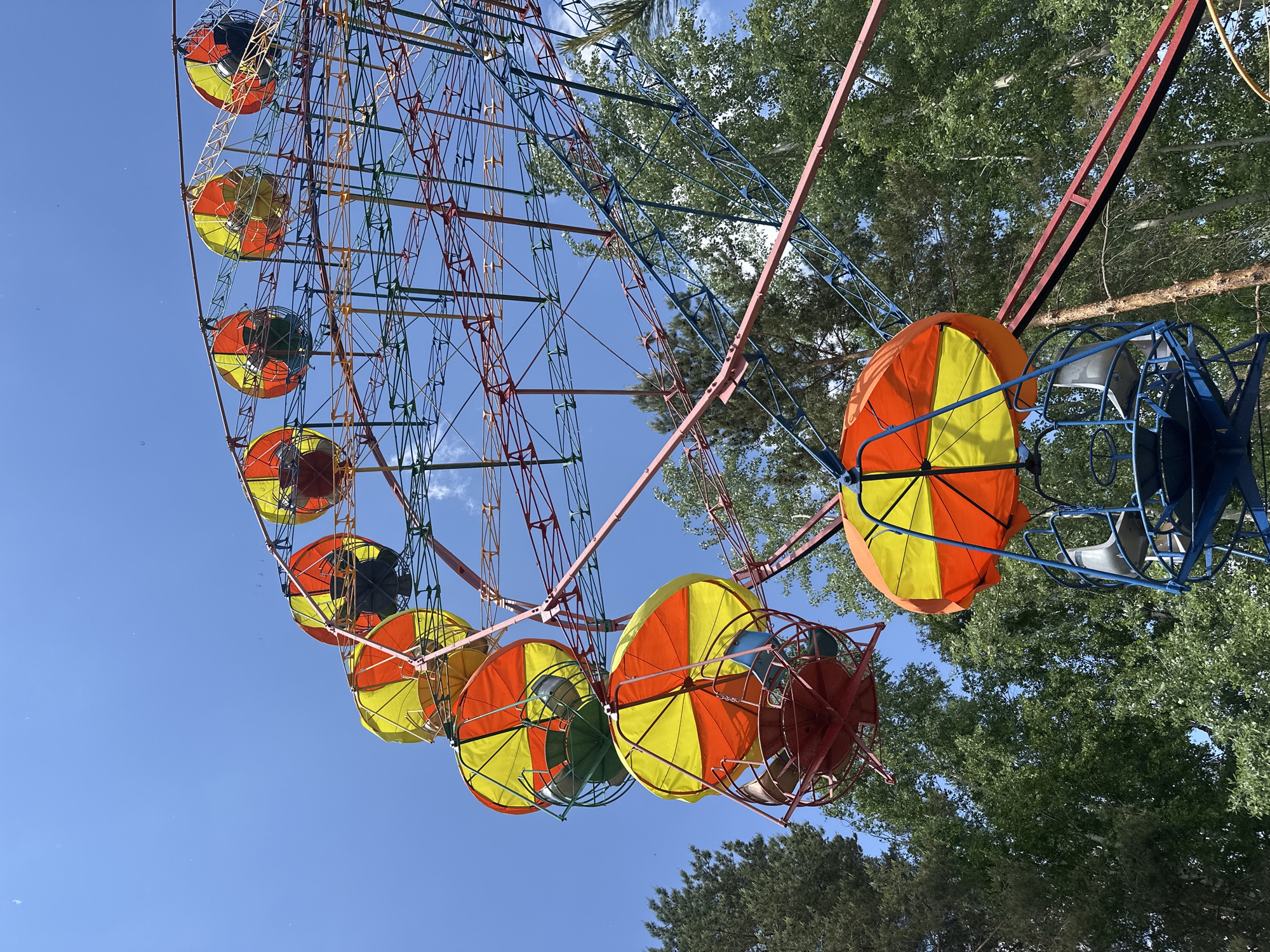 The image shows a Ferris wheel in a city park. The Ferris wheel is painted with bright colors, and the carriages are umbrella-shaped. The sky is blue, and there are trees in the background. The city is Unknown, Unknown.