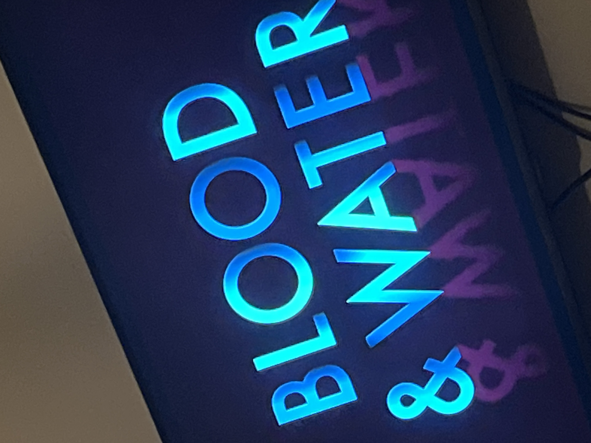 This media file is a close-up shot of a neon sign that reads "BLOOD WATER &". The letters are illuminated in a bright cyan glow against a dark purple background. The sign is angled, and the letters are slightly distorted due to the perspective. The word "BLOOD" is written vertically, with "B", "L", "O", and "O" clearly visible. Above and to the right of "BLOOD", the word "WATER" is also written vertically, with "W", "A", "T", "E", and "R" visible. The ampersand symbol "&" is positioned below and to the right of the word "WATER". Faint pink and purple shadows of the letters are visible behind the neon glow, suggesting that the sign might be casting a reflection or that there is another similar sign behind it. The overall impression is one of a vibrant, possibly nocturnal, display. No people are visible in the image. The setting appears to be indoors or a covered outdoor space, given the consistent lighting and the presence of what looks like wires or cables on the right side of the image, possibly connected to the sign. The image is likely taken at night or in a dimly lit environment to highlight the neon illumination. There are no discernible weather cues. The location context provided is Gura Top, Nigeria.