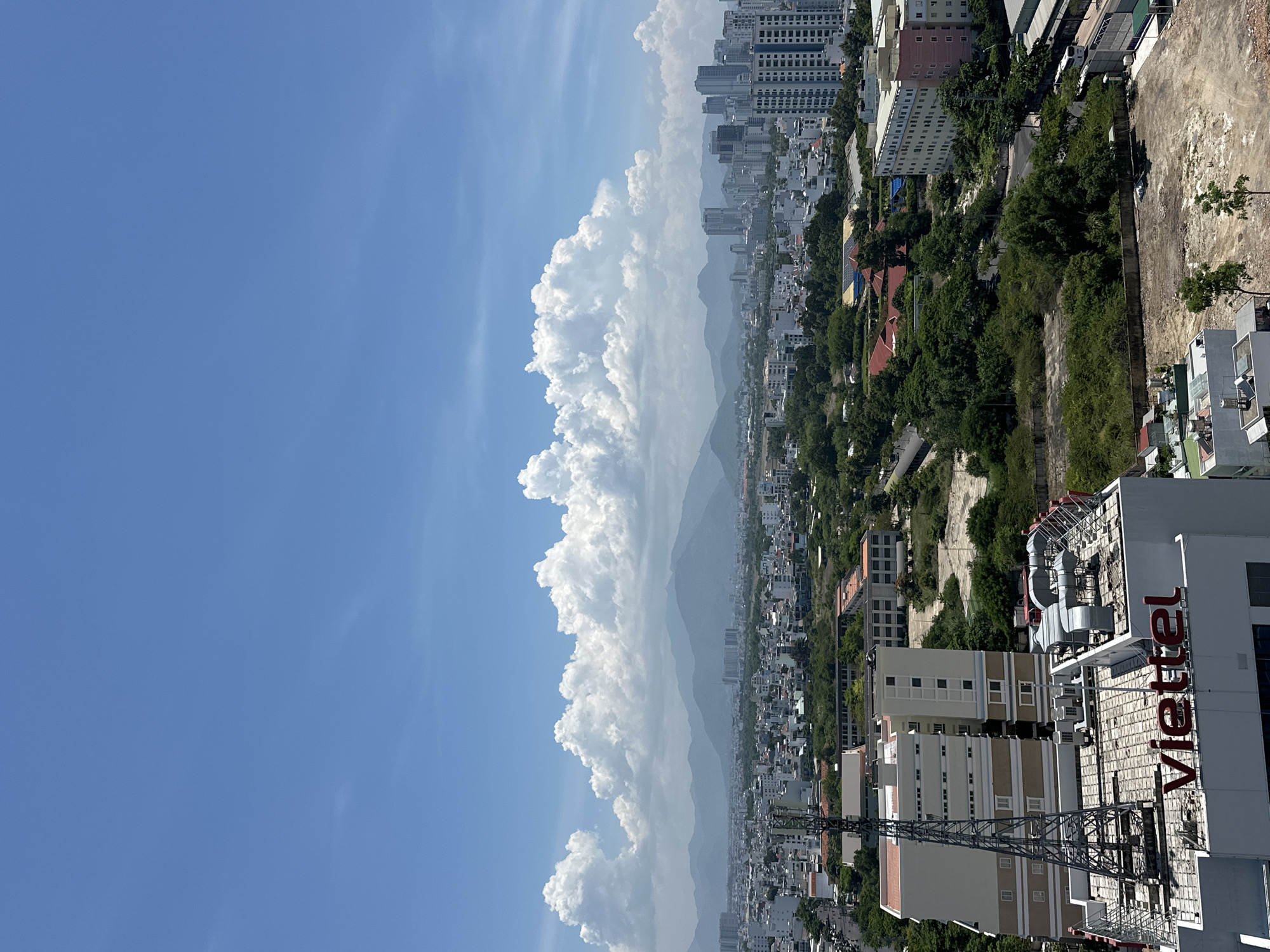 This is a high-angle, wide shot of a city skyline under a bright blue sky with scattered white clouds. The foreground features a large building with a prominent "viettel" logo in red letters on its roof. A tall, metal tower, likely a communication mast, stands beside this building. Beyond this, the city stretches out with a mix of mid-rise and high-rise buildings, interspersed with patches of green trees and vegetation. In the distance, a range of hazy mountains forms the backdrop. The overall impression is a sunny day in a developed urban area. The location is identified as Nam Nha Trang Ward, Vietnam. There are no people visible in the image.