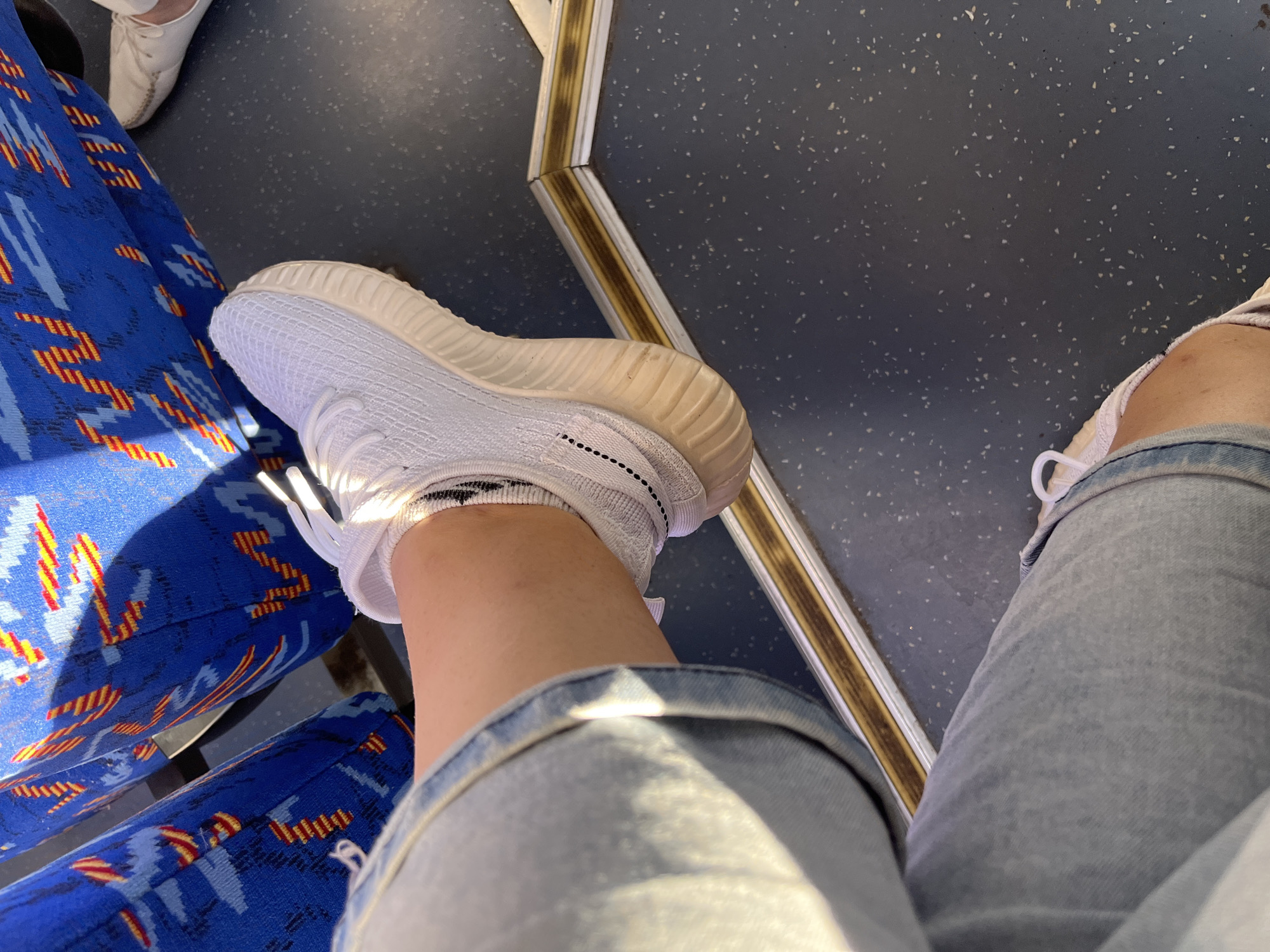 A person's leg, clad in light-wash denim jeans and a white sneaker with black trim, is positioned on a bus seat. The seat fabric is a vibrant blue with an abstract red and yellow pattern. The bus floor is dark gray with light speckles. A metallic trim with a gold and brown stripe runs along the floor. The scene is illuminated by natural sunlight, casting shadows. The location is identified as a bus in Myadzyel, Belarus.